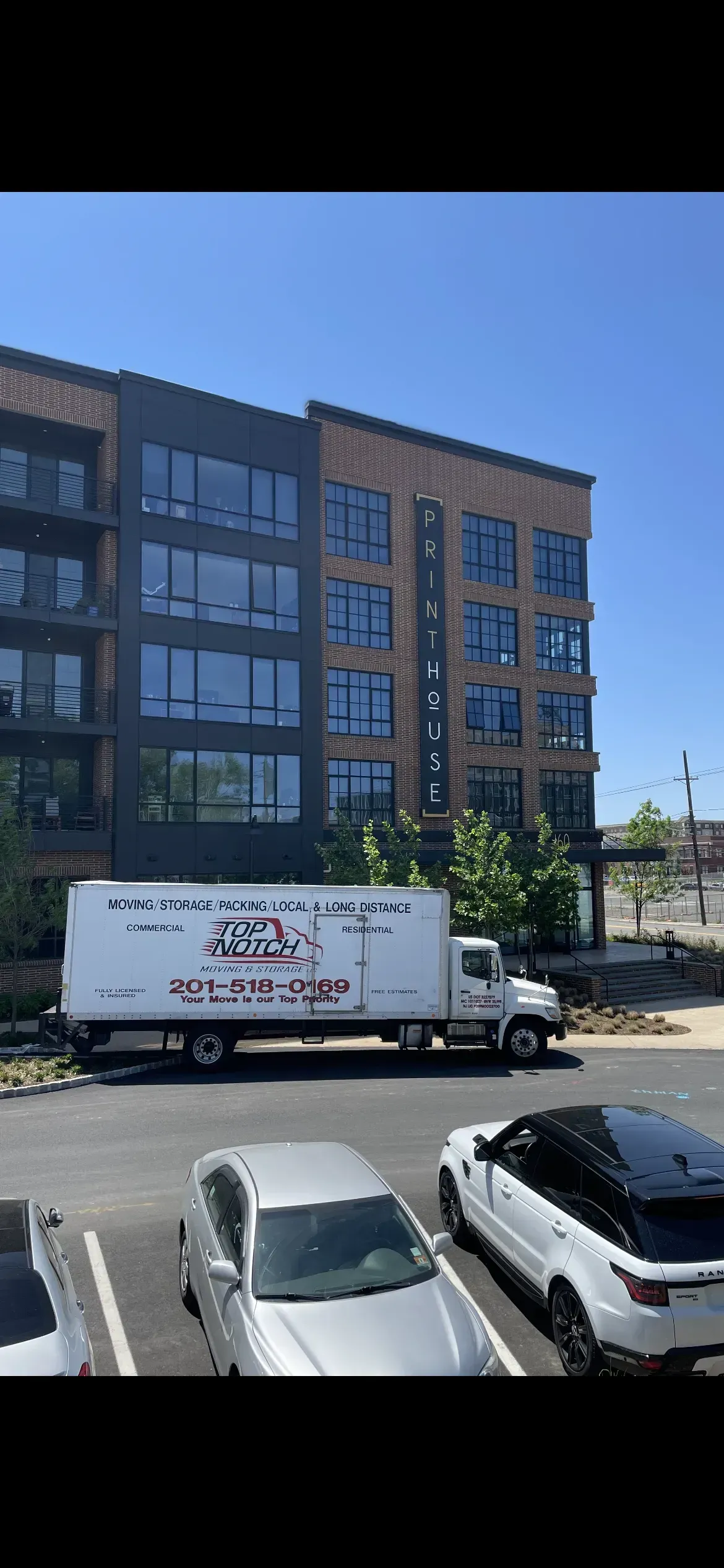A moving truck parked in front of an apartment building on a sunny day. Cars are parked nearby.