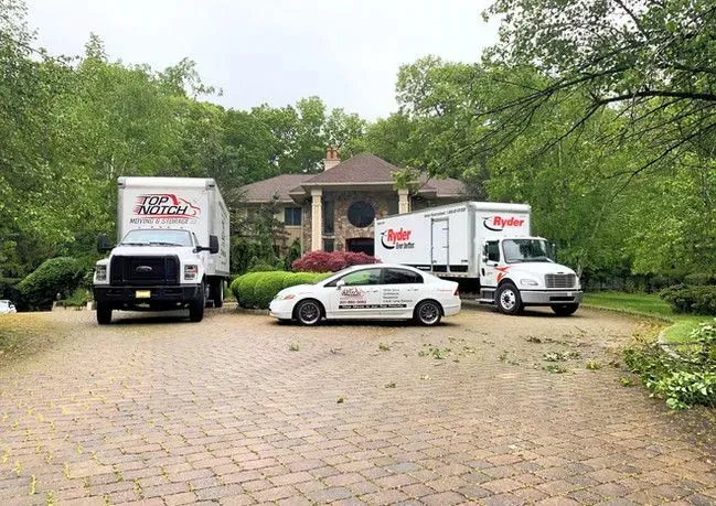 Two moving trucks and a car parked in front of a house. Paved driveway, green trees.