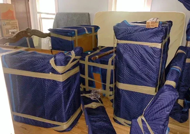 Furniture wrapped in blue moving blankets, secured with tan tape, ready for transport.