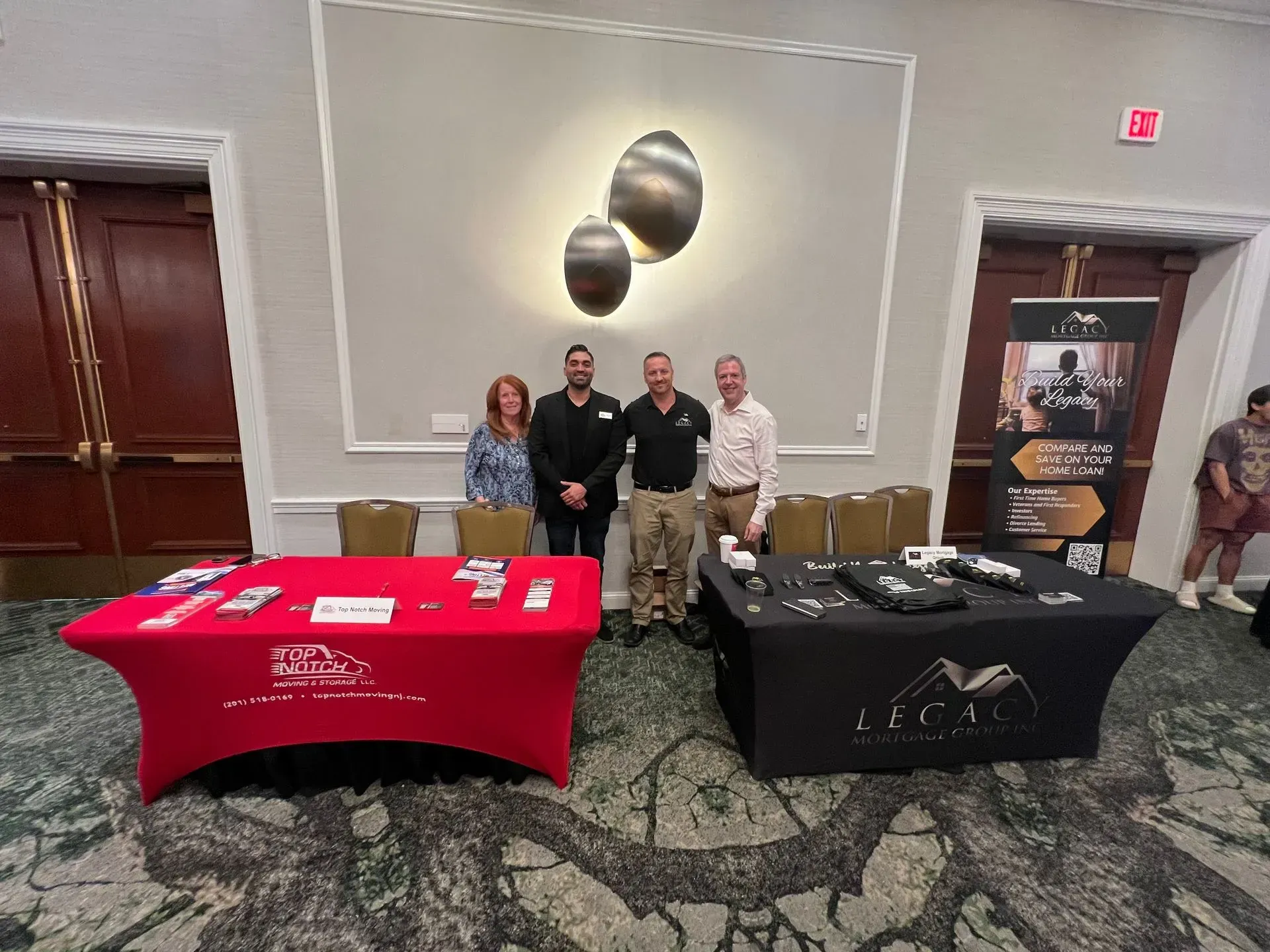 People stand behind tables displaying products at a conference. Two tables have company logos. A banner stands on the right.