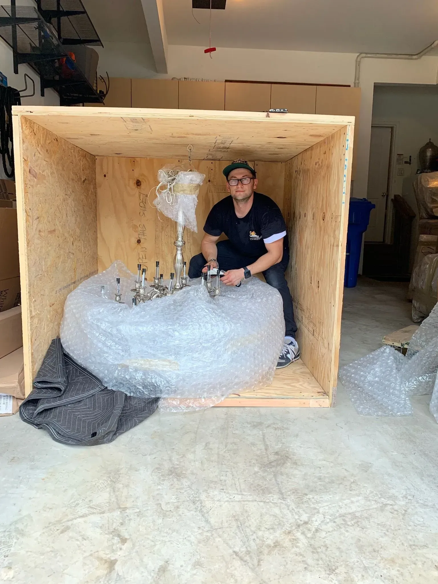 Man in a wooden crate with a chandelier wrapped in bubble wrap; in a garage.