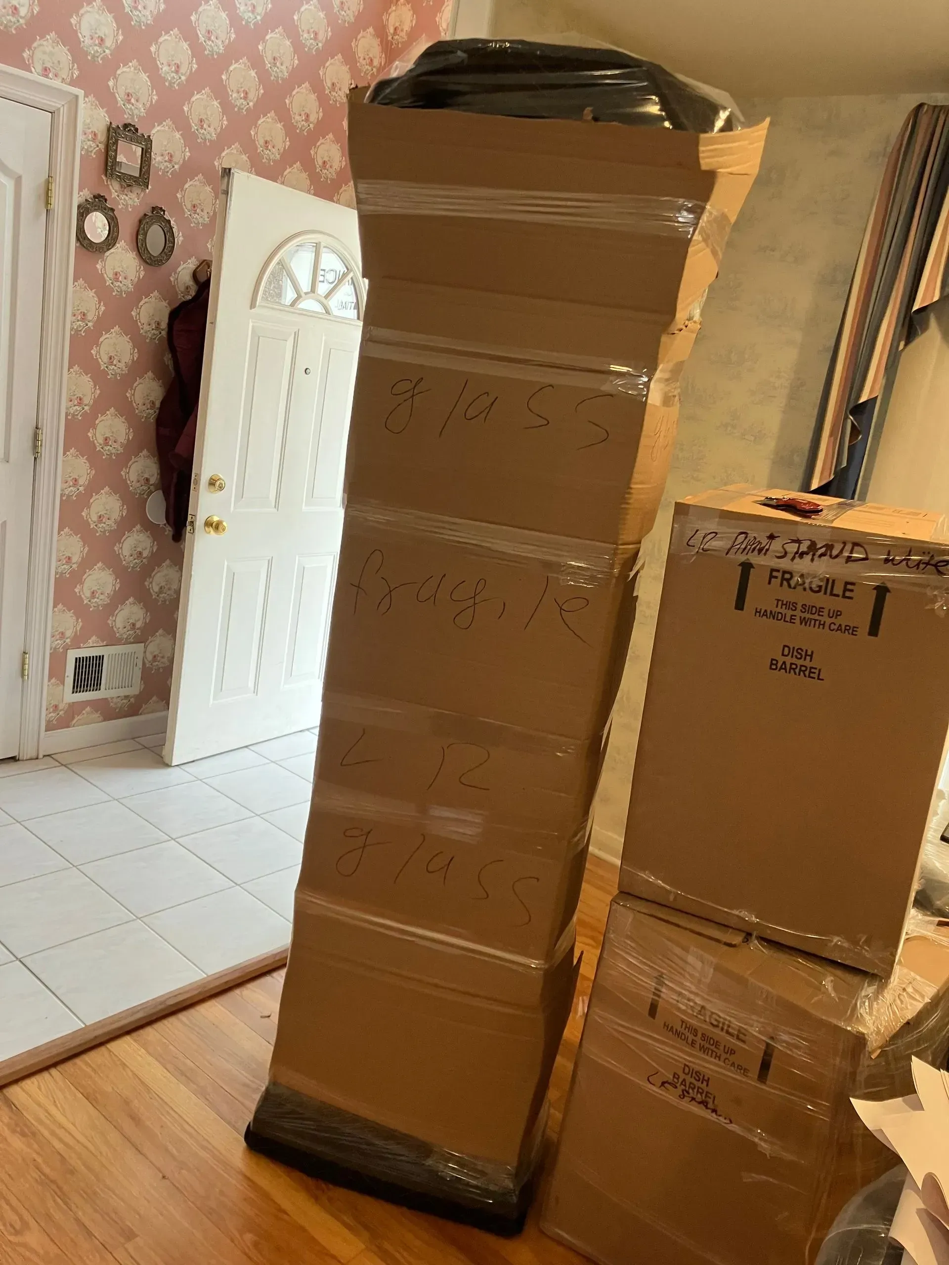 Tall, wrapped cardboard package leans against other boxes near a doorway; pink wallpaper in the background.