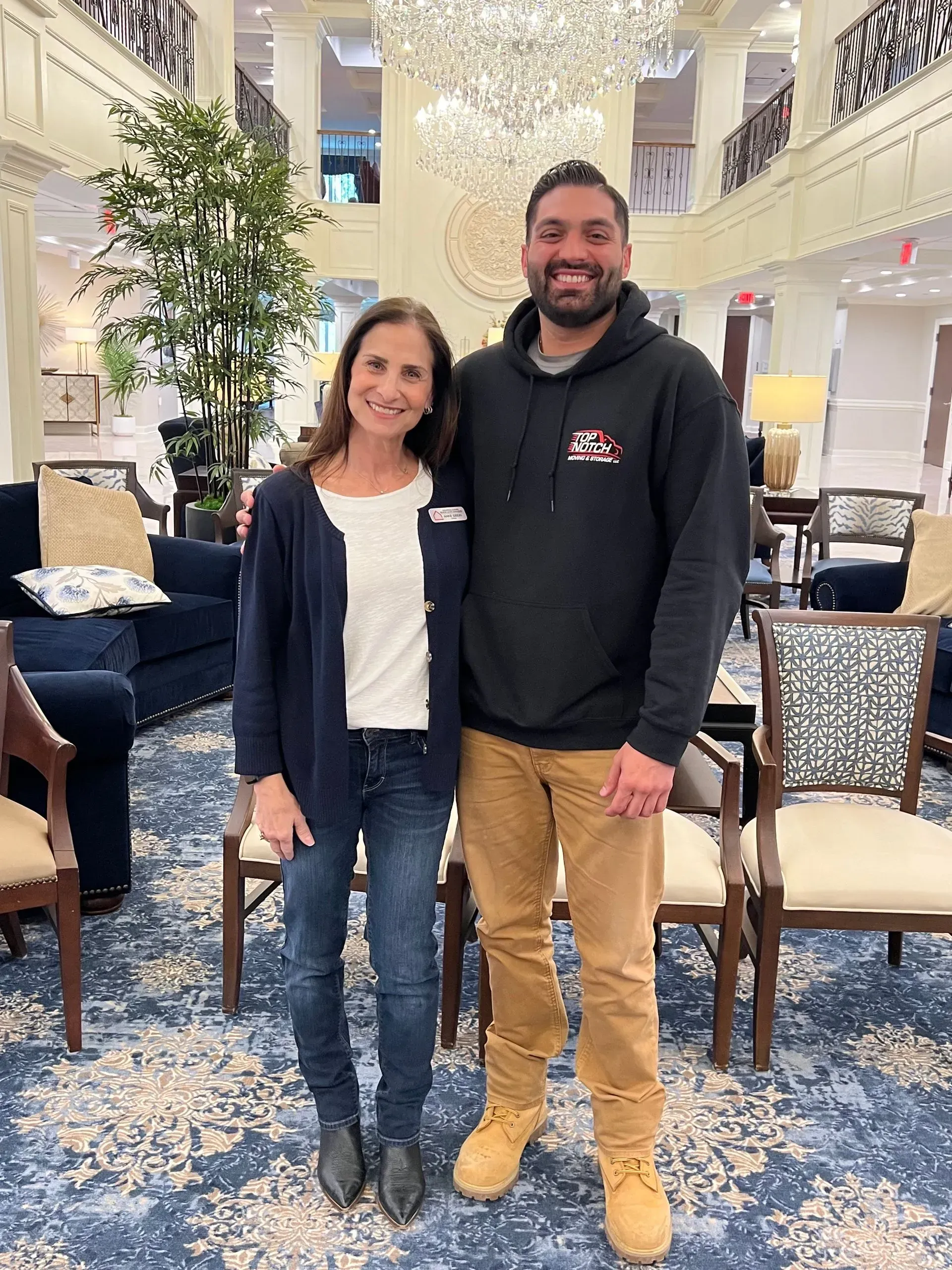 Woman and man standing in an ornate lobby. Man wears a black hoodie and tan pants; woman wears a cardigan and jeans.