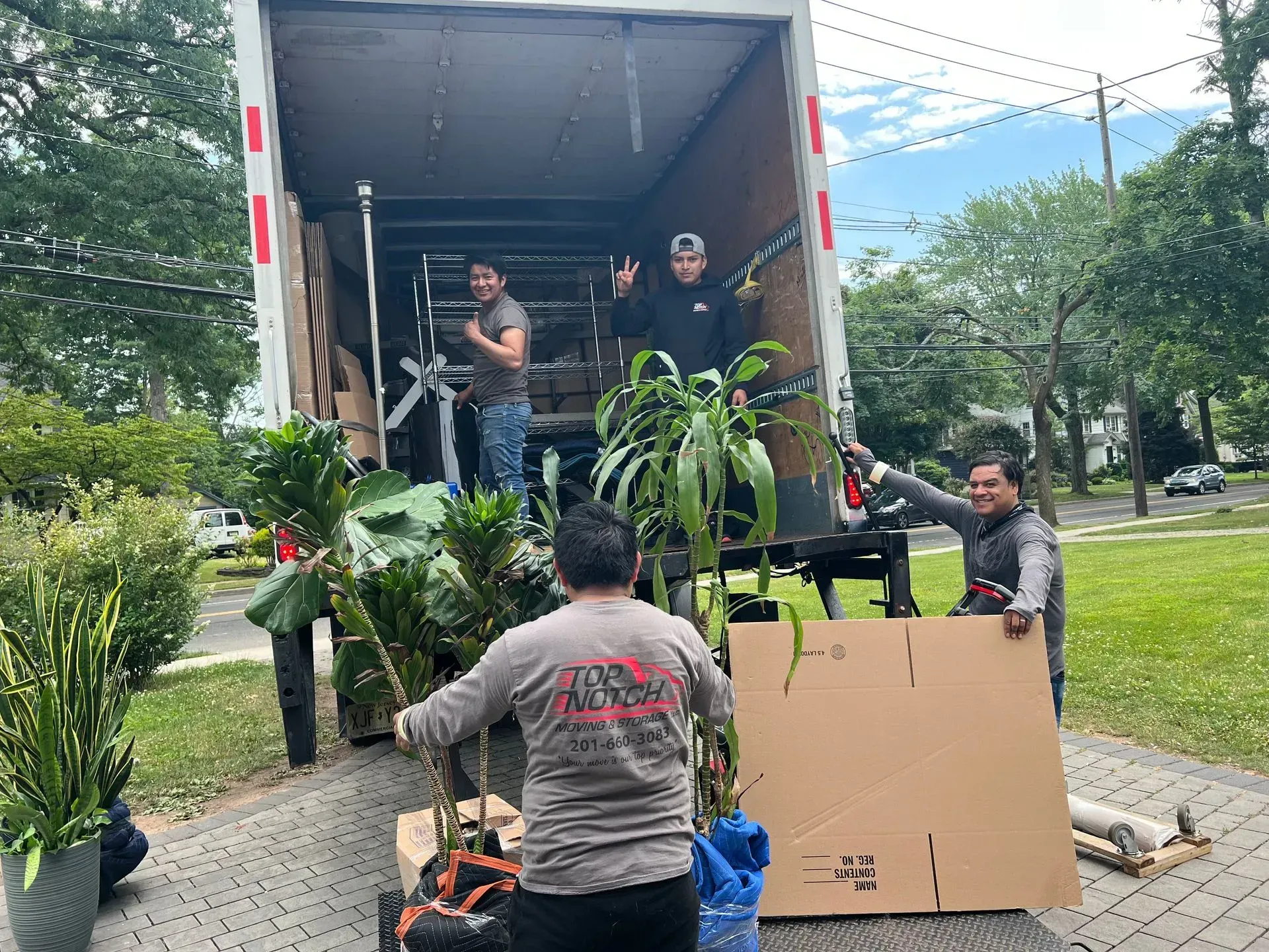 Movers loading a truck with plants, a cardboard box, and furniture on a residential street.