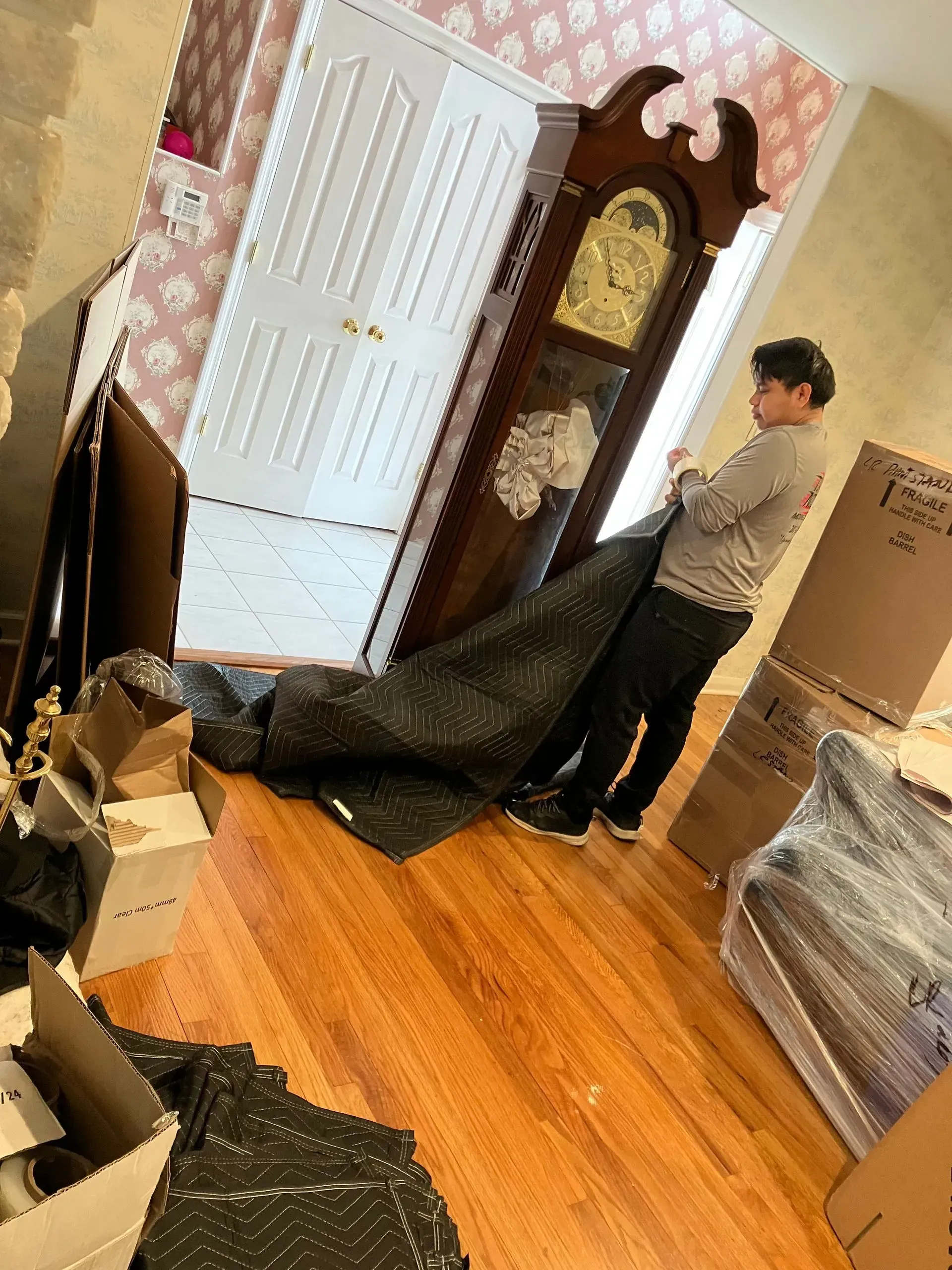 Person wrapping a tall, wooden grandfather clock with moving blankets near a doorway. Boxes on the floor.