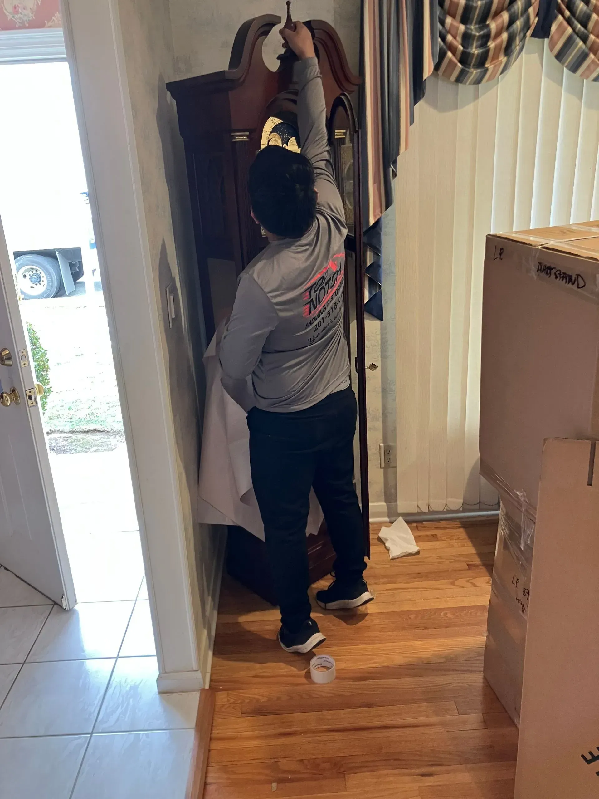 Person winding a grandfather clock in a room with wood floors and moving boxes.