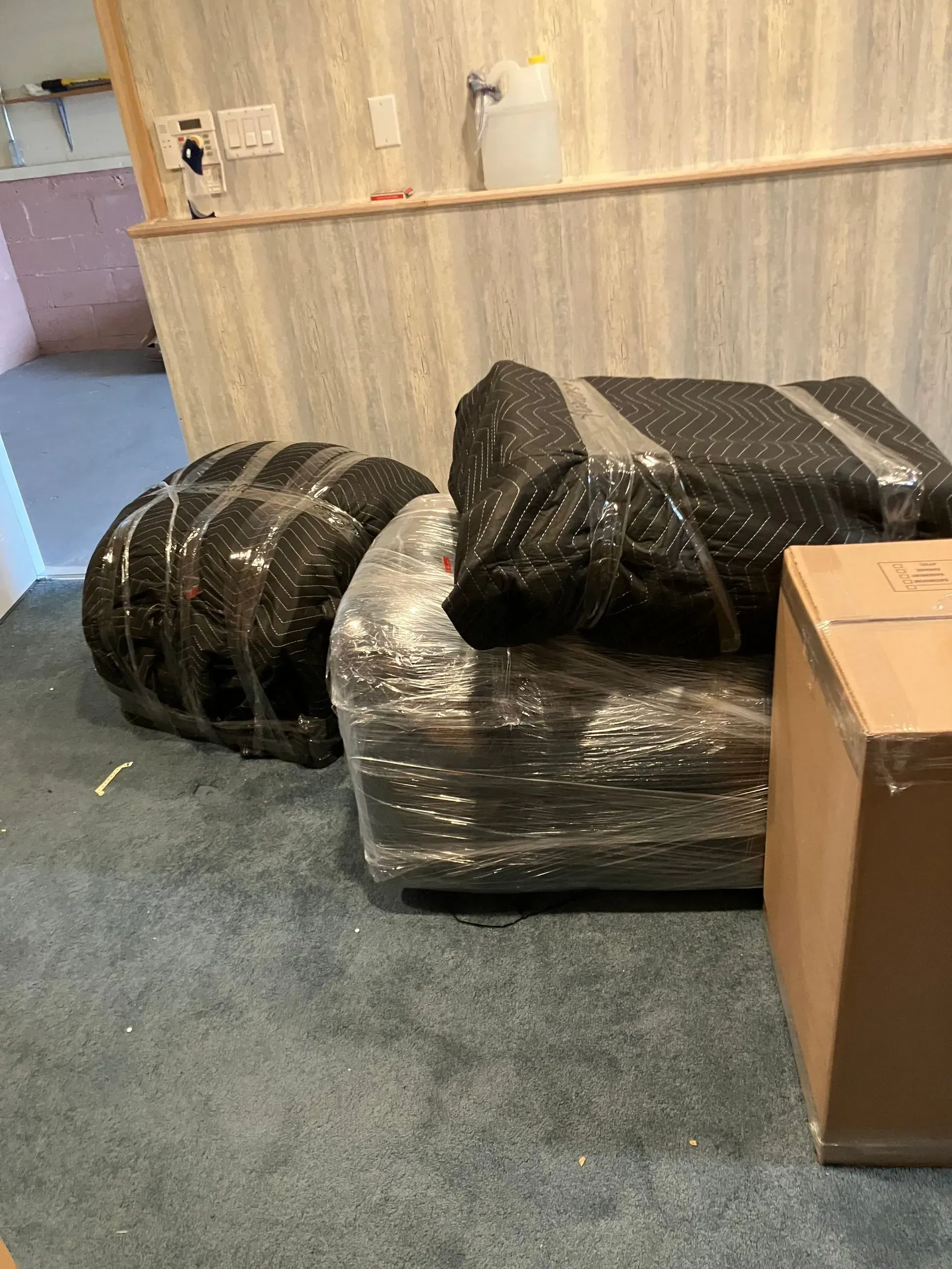 Furniture wrapped in black plastic and clear wrap; cardboard box on blue carpet.