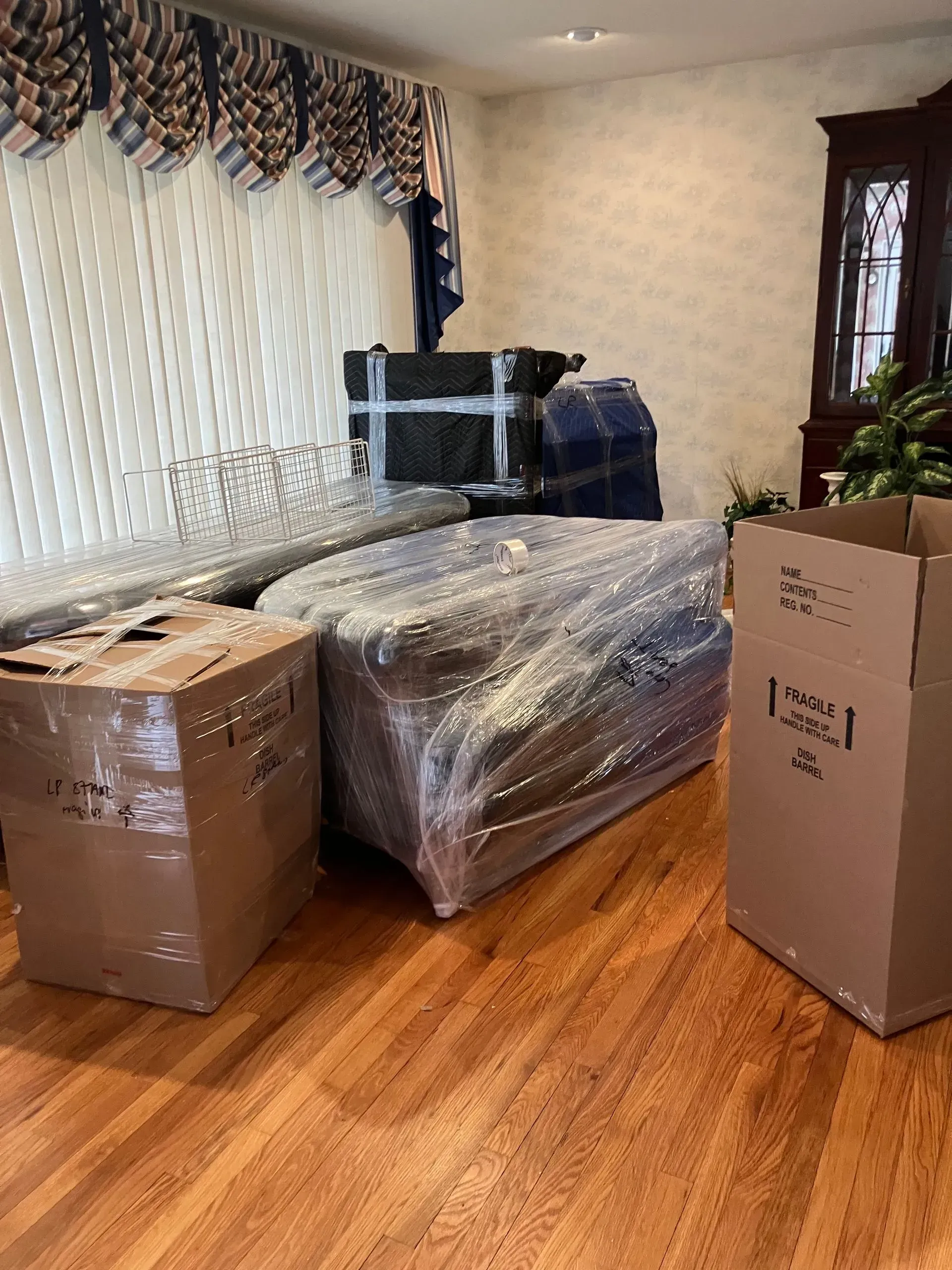 Boxes and furniture wrapped in plastic and tape in a room with hardwood floors, curtains, and a window.