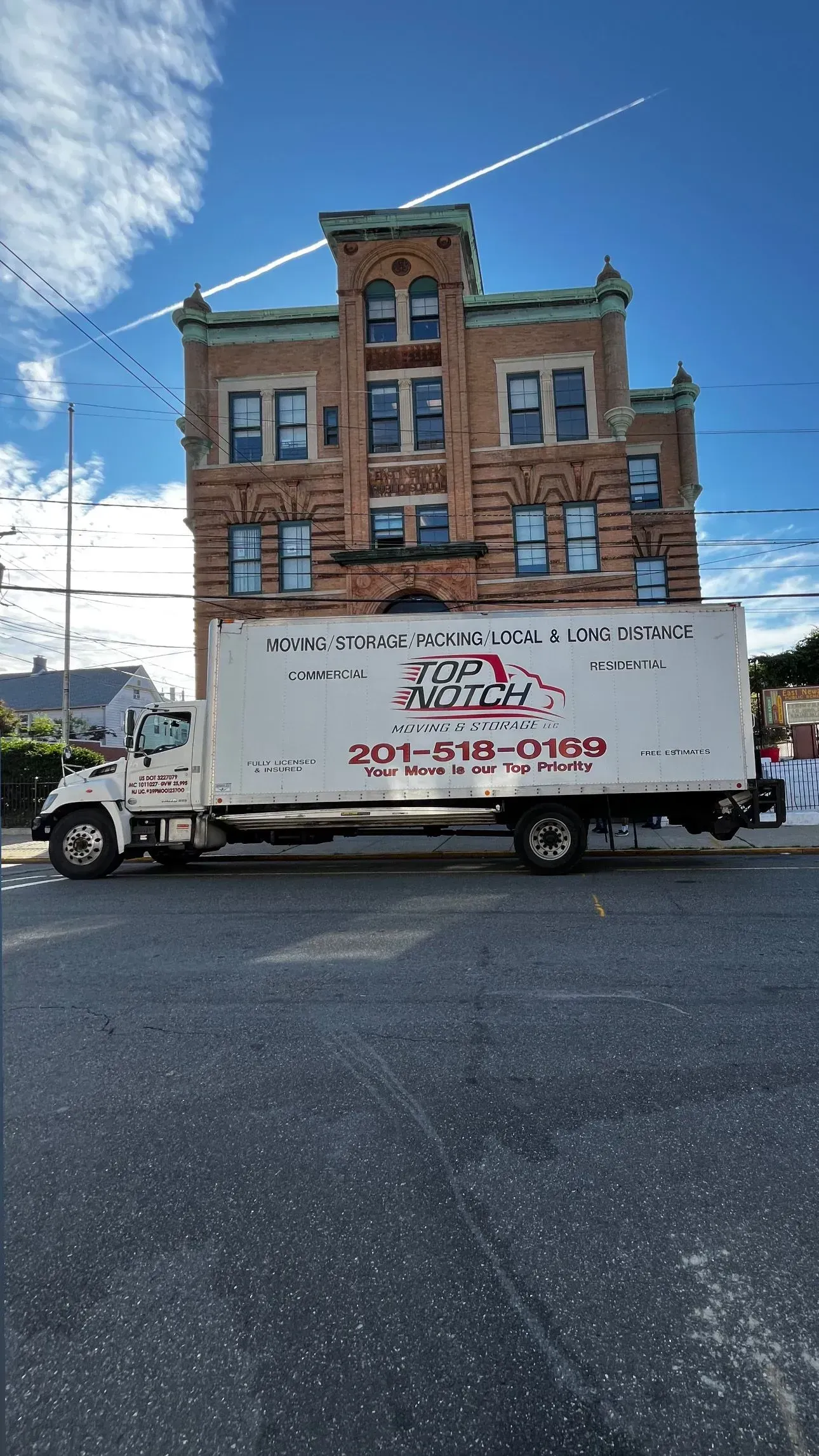 Moving truck parked in front of a brick building. Truck has company logo; building is several stories high. Blue sky overhead.