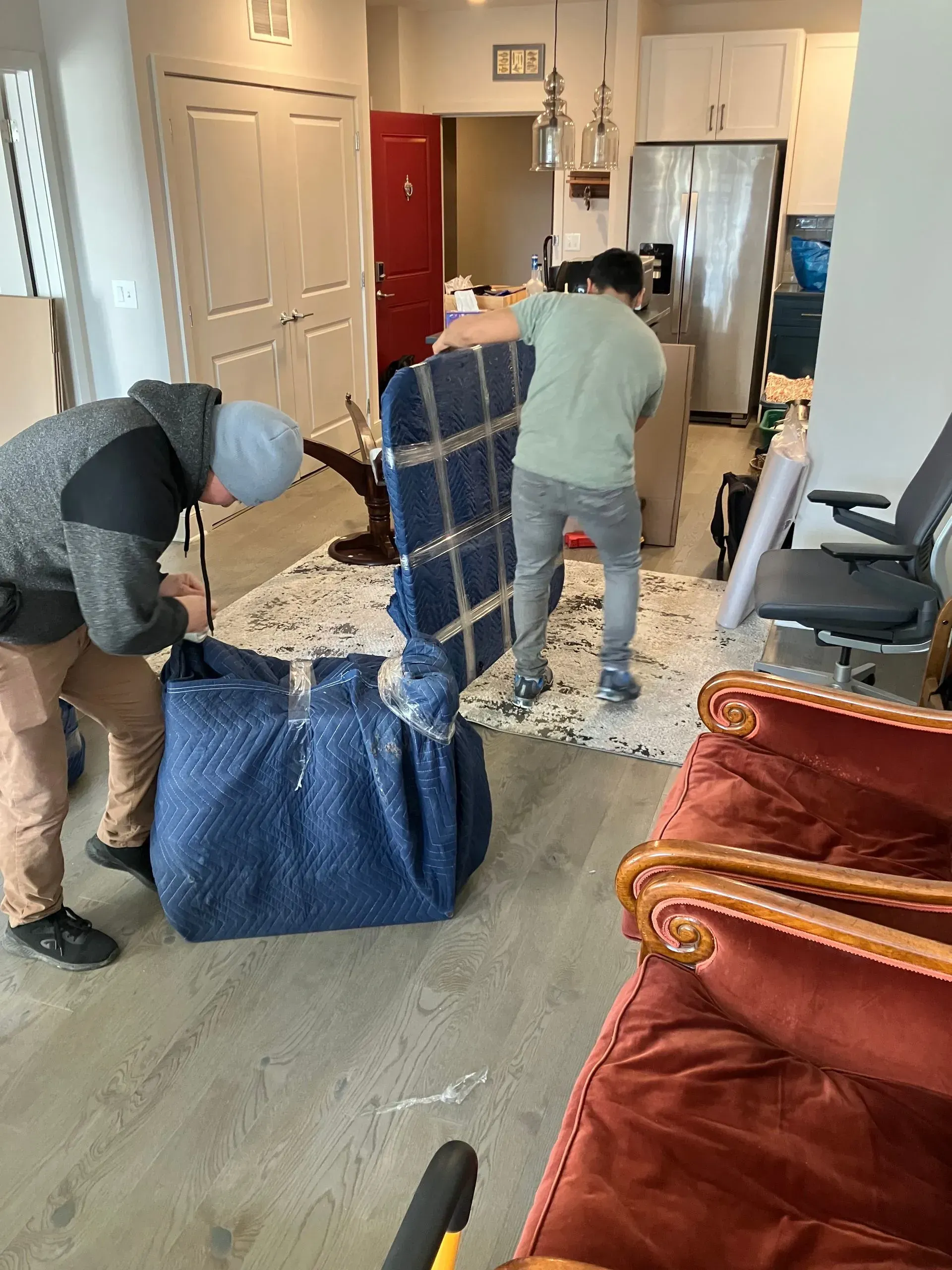 Two people packing furniture indoors; one holds a wrapped piece, another unrolls padding, debris on floor.