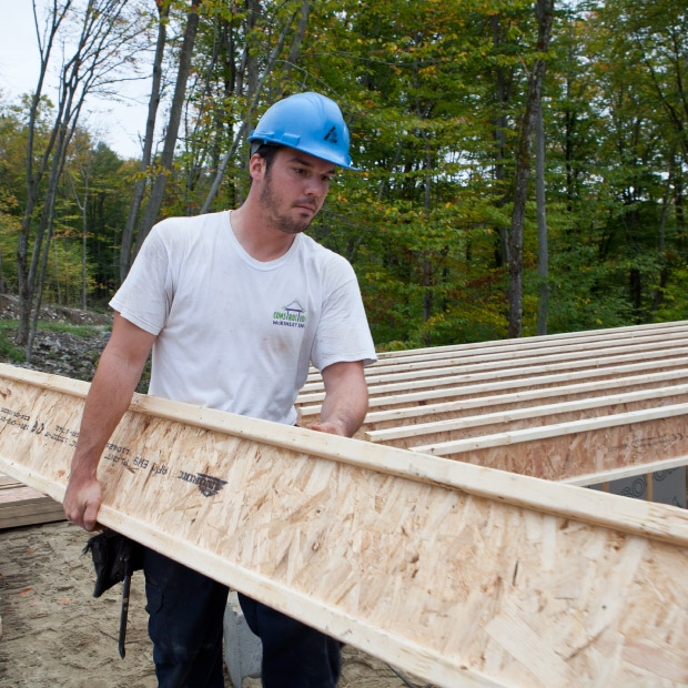 Construction worker in a blue helmet carrying a long wooden beam at a construction site.