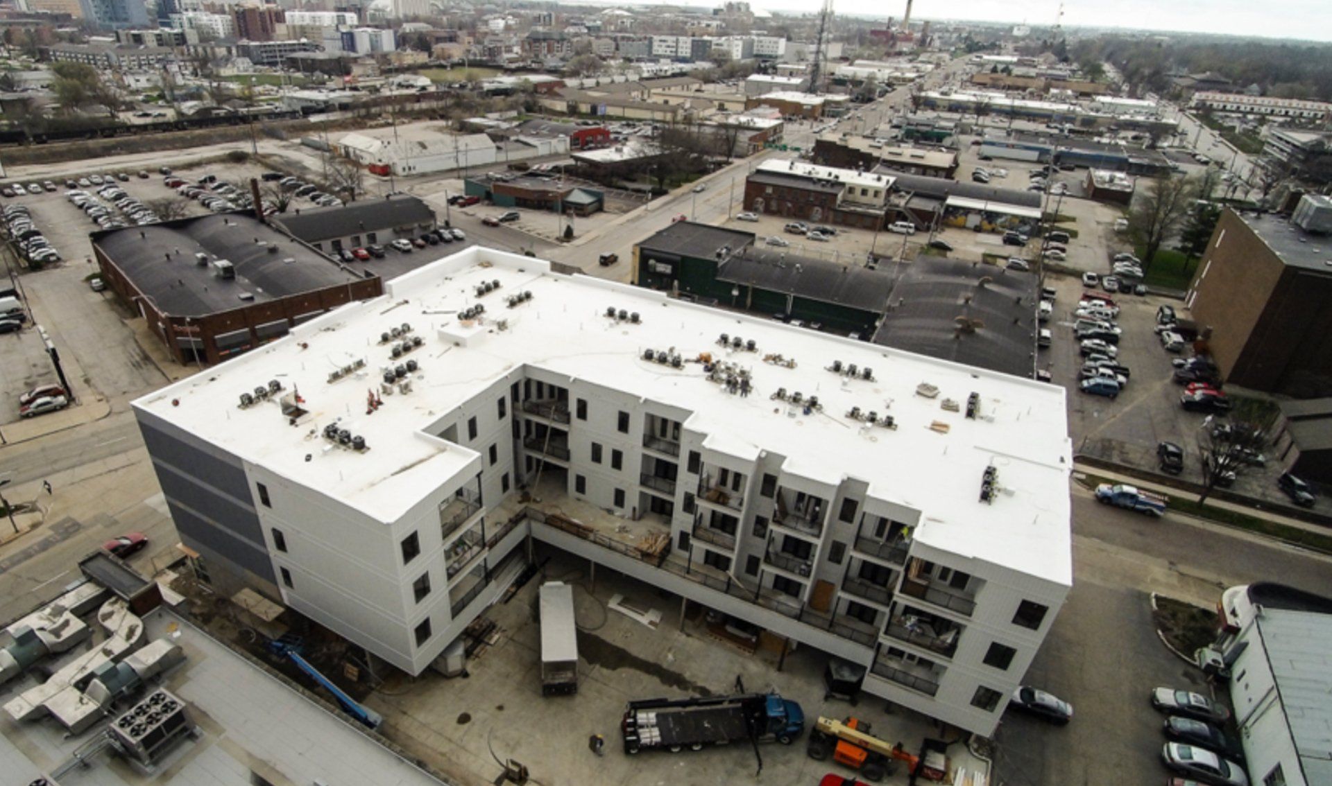Aerial view of a modern, white L-shaped apartment building with balconies, surrounded by city buildings and parking lots.