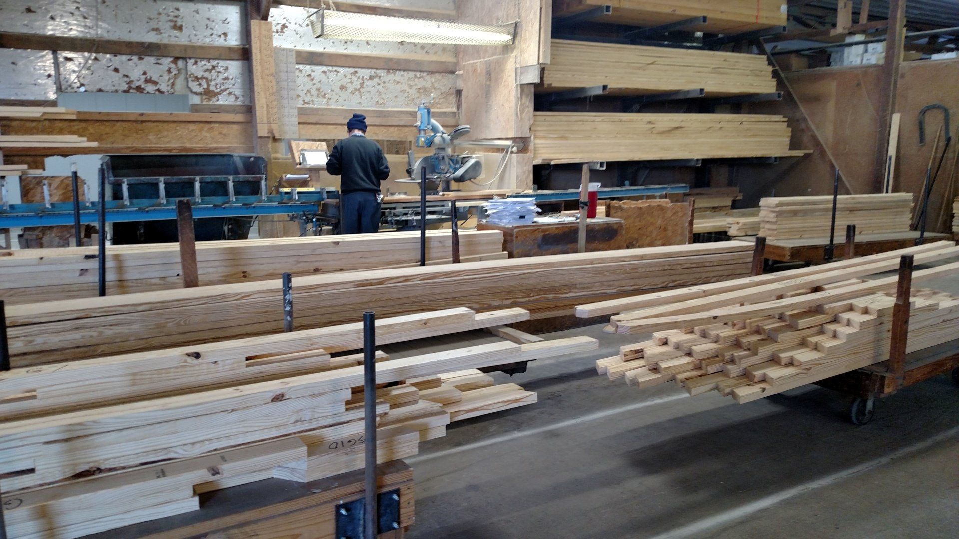Lumber yard with worker operating machinery. Rows of wooden beams on carts and shelves, indoors.