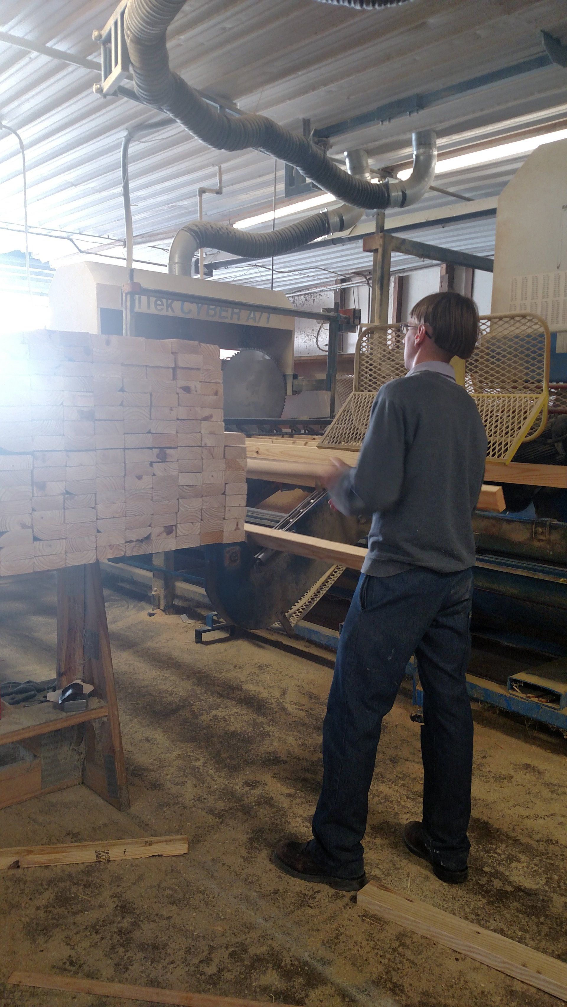 Person in a wood workshop operating machinery, handling wood, sawdust on the floor, overhead ventilation.