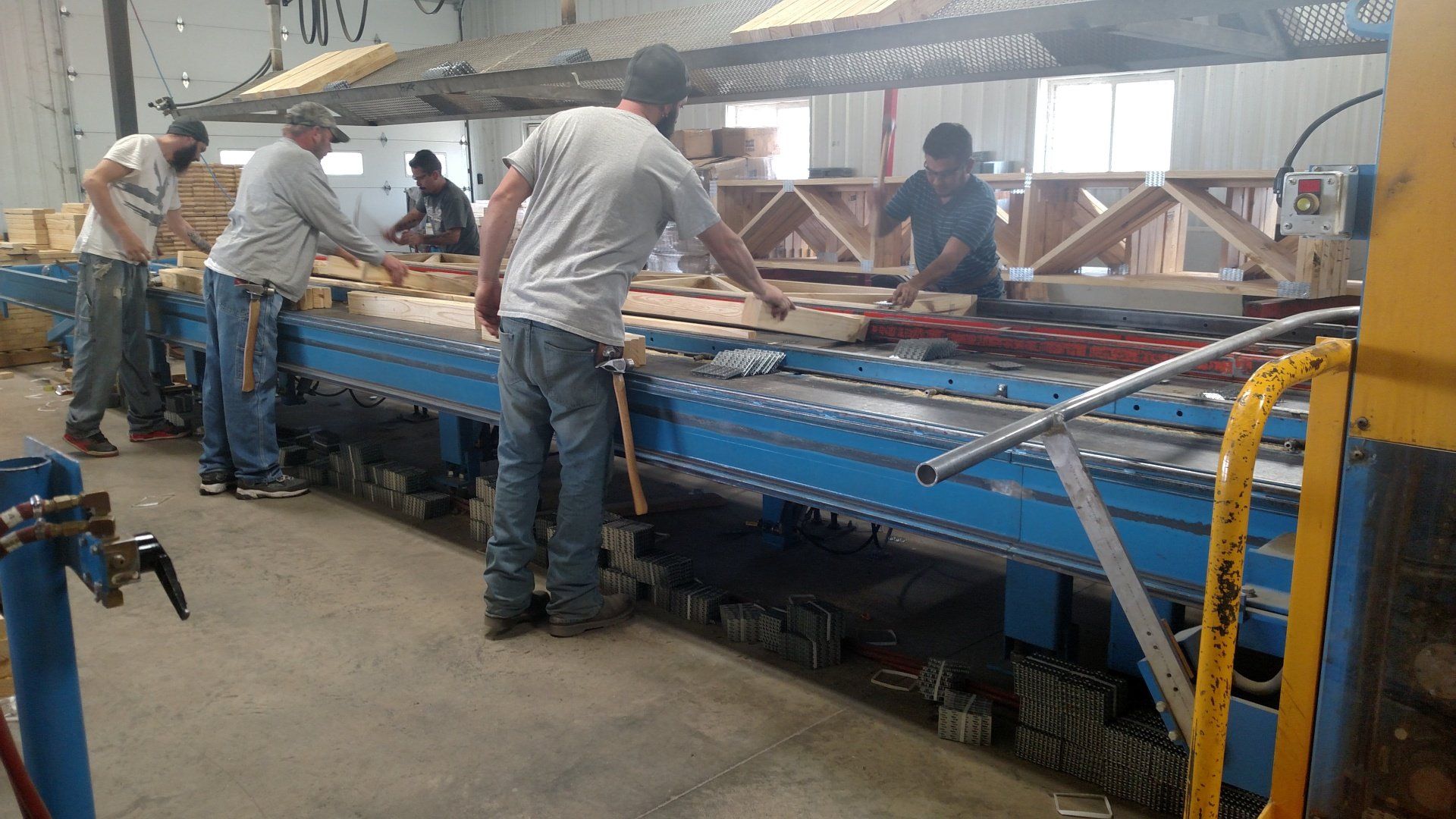 Workers in a factory assembling wooden trusses on a blue conveyor belt.
