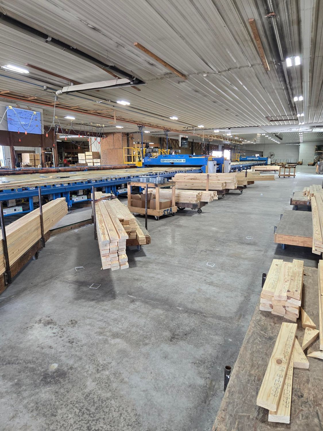 Lumber mill interior with stacks of wood on conveyor belts. Industrial ceiling and concrete floor.