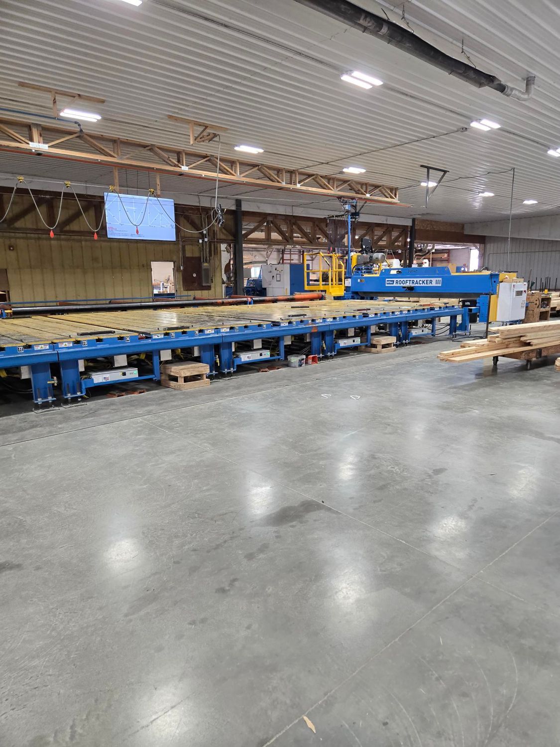 Large blue industrial machine in a factory setting, processing wooden boards, with a bright overhead lighting system.