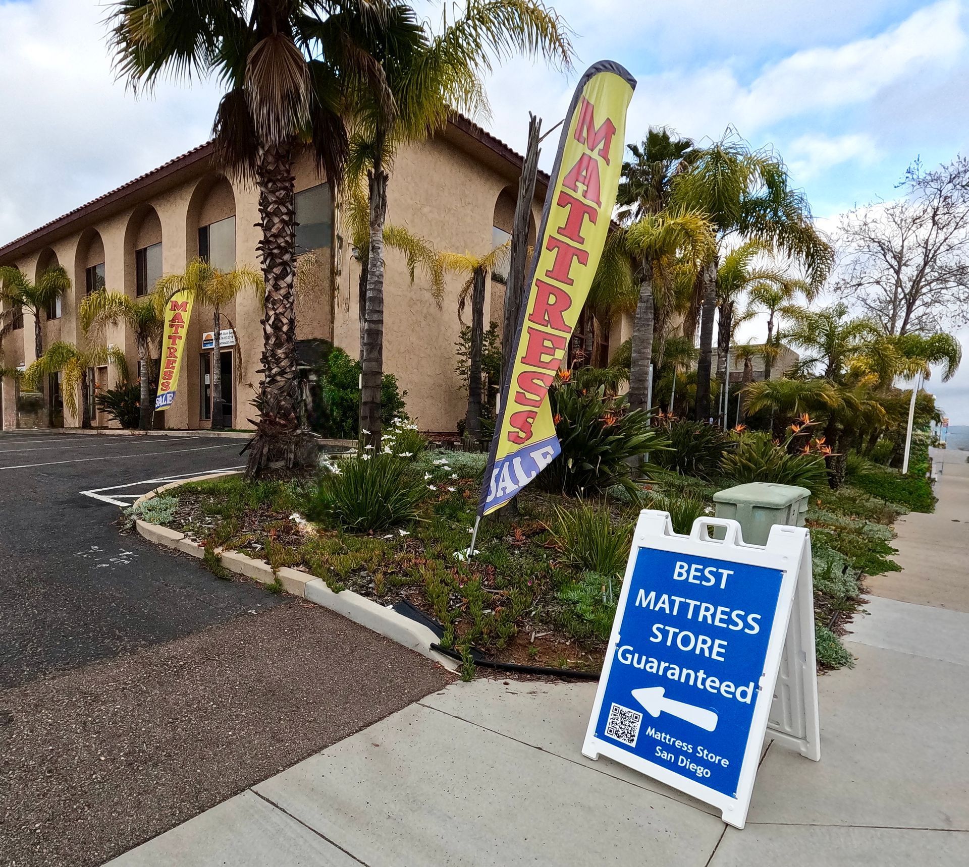 Exterior of a mattress store with signs: 'Mattress' and 'Best Mattress Store Guaranteed'. Beige building, palm trees.