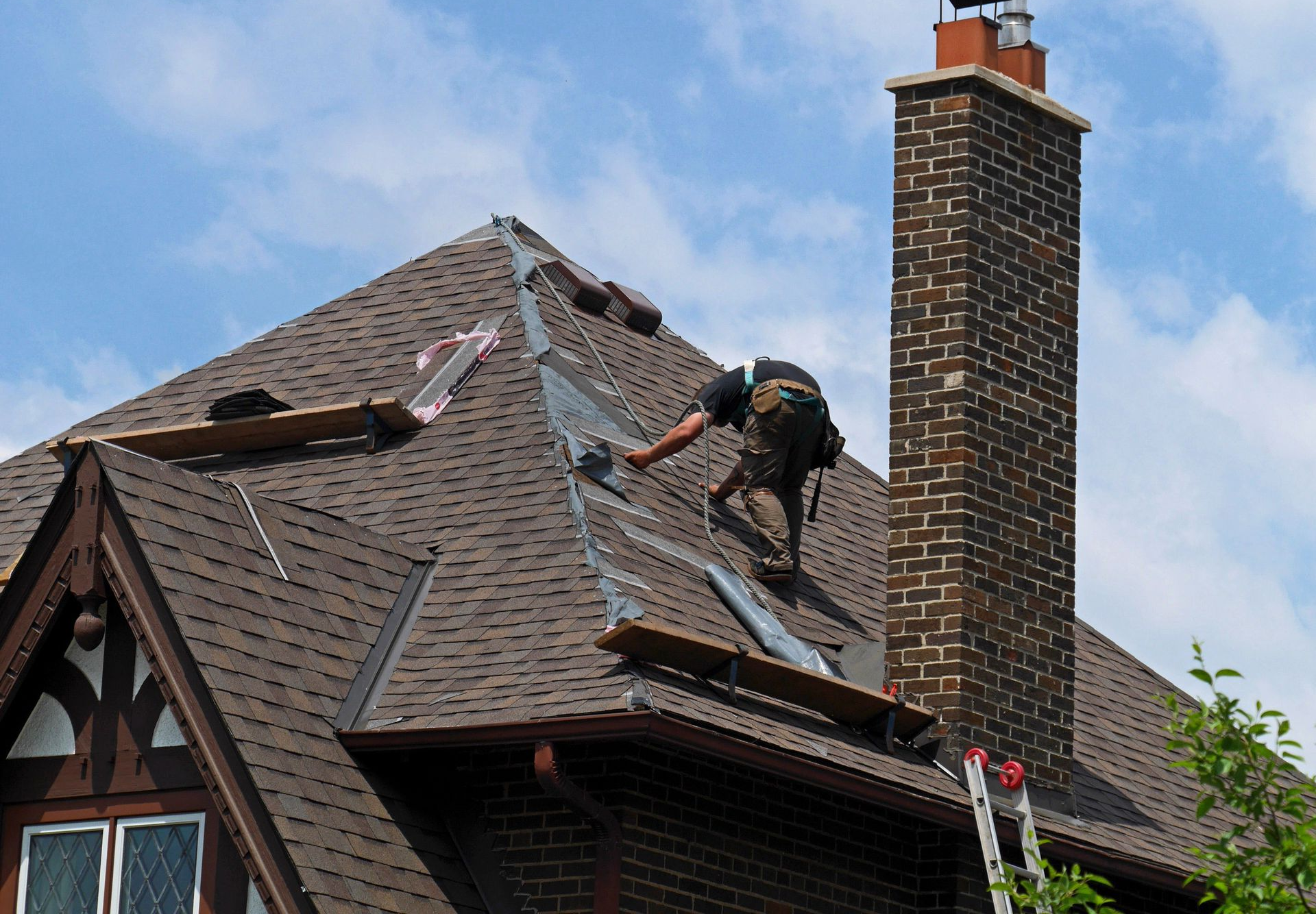 Roofers working on a steep brown shingle roof beside a brick chimney under a blue sky