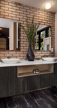 Modern bathroom vanity with double sinks, mirrors, brick wall, and a tall vase of greenery