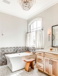 Bright bathroom with freestanding tub, wood vanity, patterned tile, and chandelier near a window.