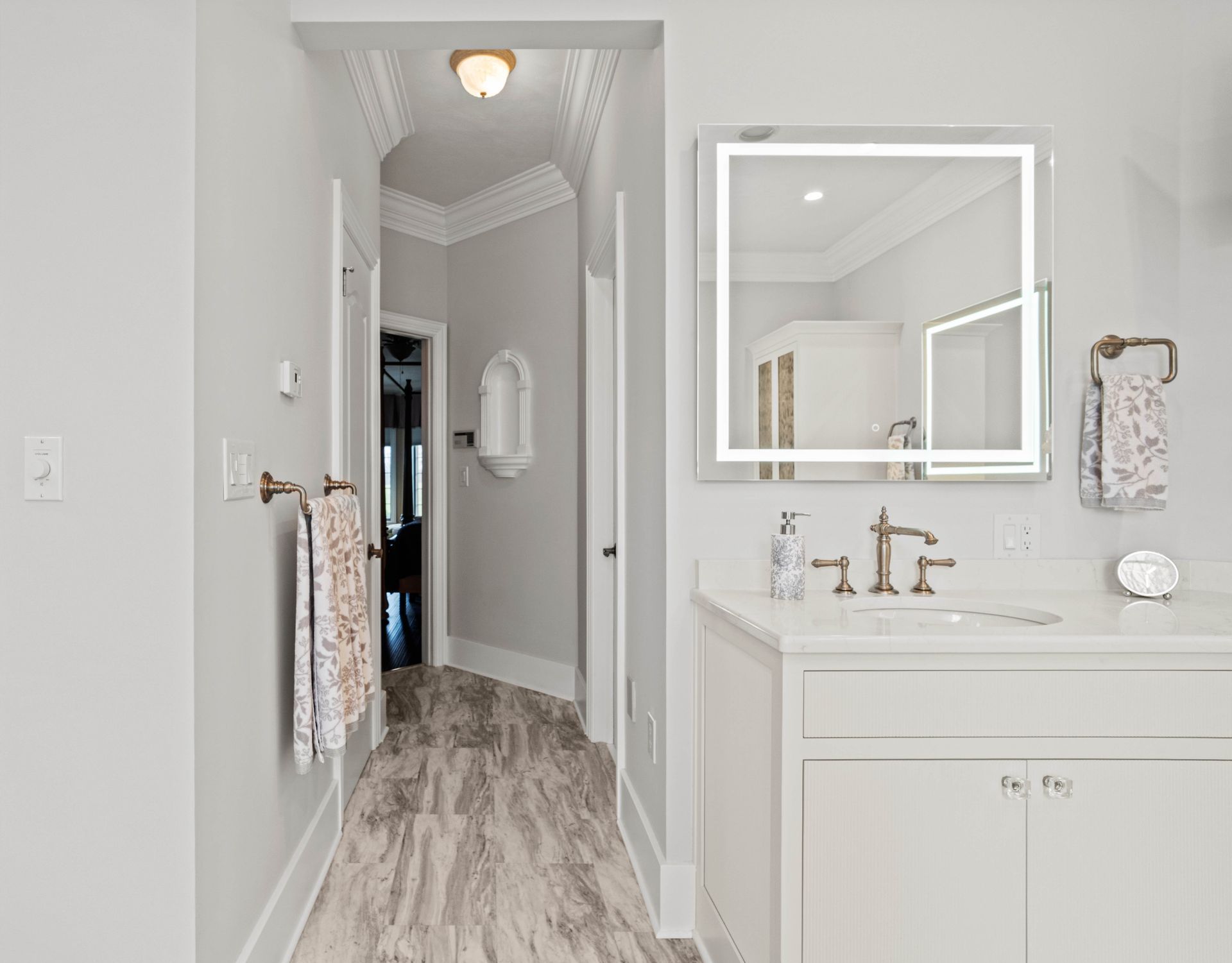 Bright bathroom hallway with white vanity, mirror, and light wood floors leading to a doorway.
