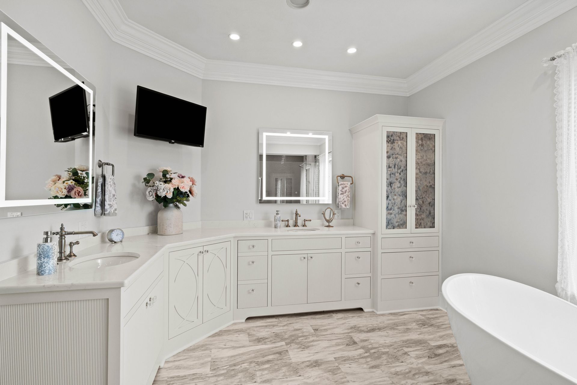 Bright white bathroom with double vanity, wall-mounted TVs, large mirror, and freestanding tub