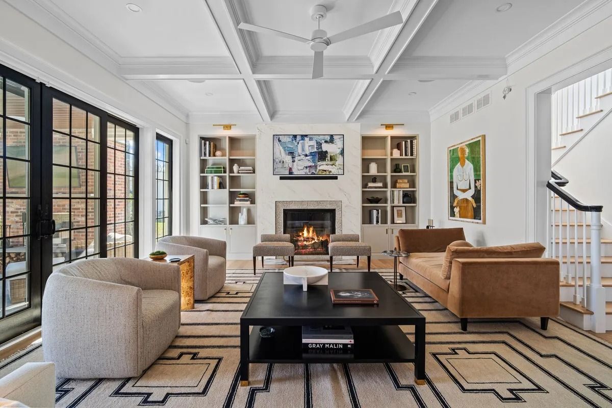 Bright modern living room with fireplace, built-in shelves, tan and white seating, and large black-framed windows.