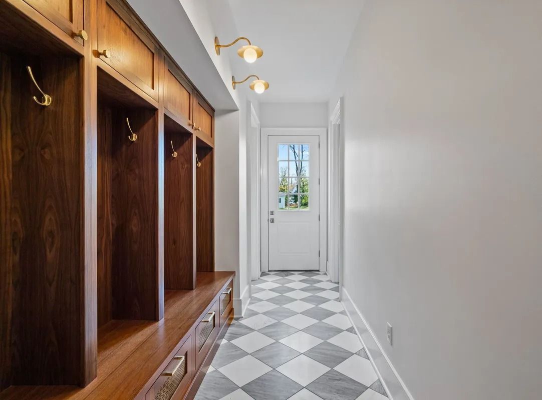 Narrow hallway with wooden built-in cubbies, white walls, and patterned black-and-white tile floor leading to a door.