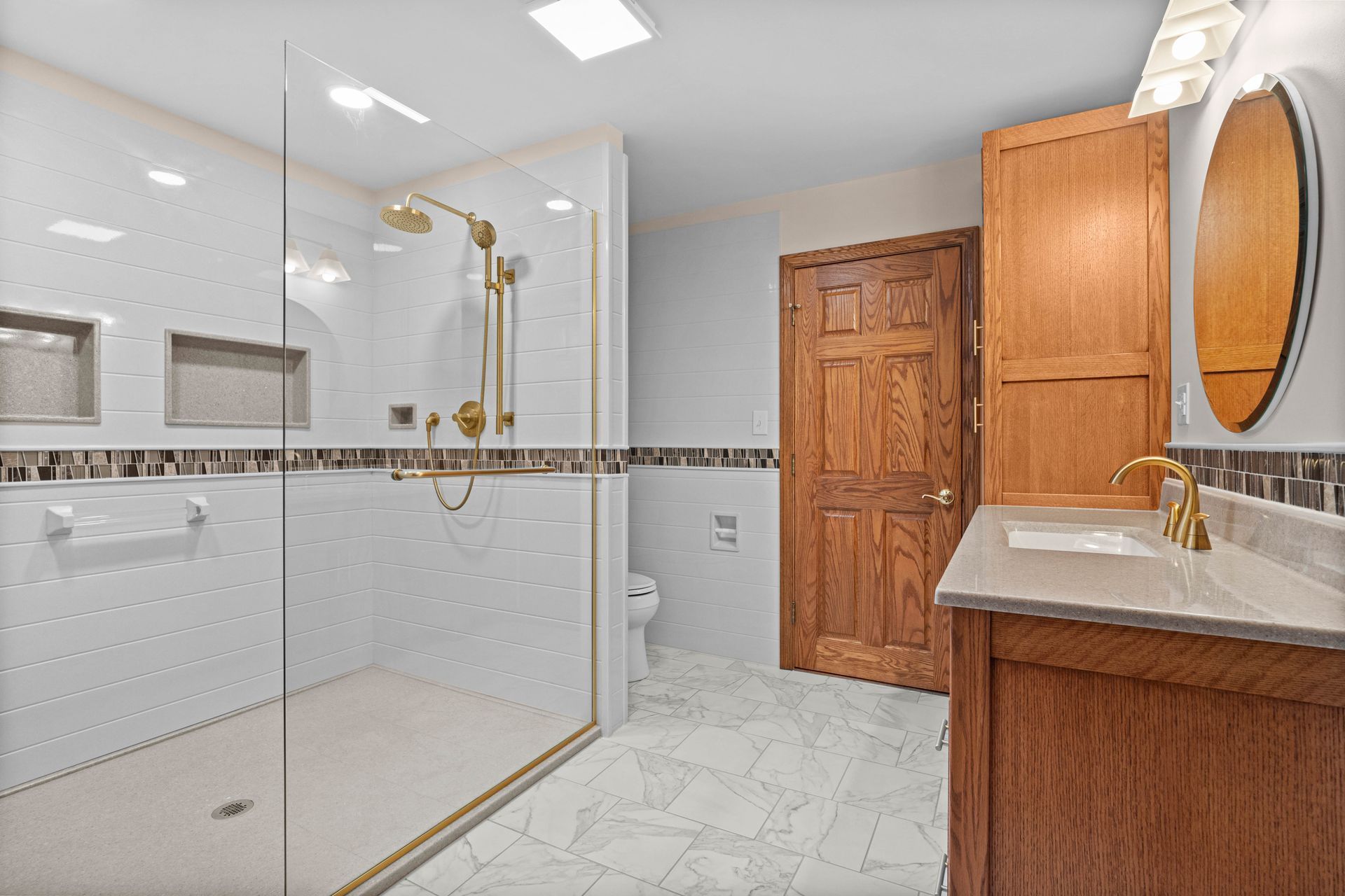 Bright modern bathroom with glass shower, white tile, and wood vanity with oval mirror.