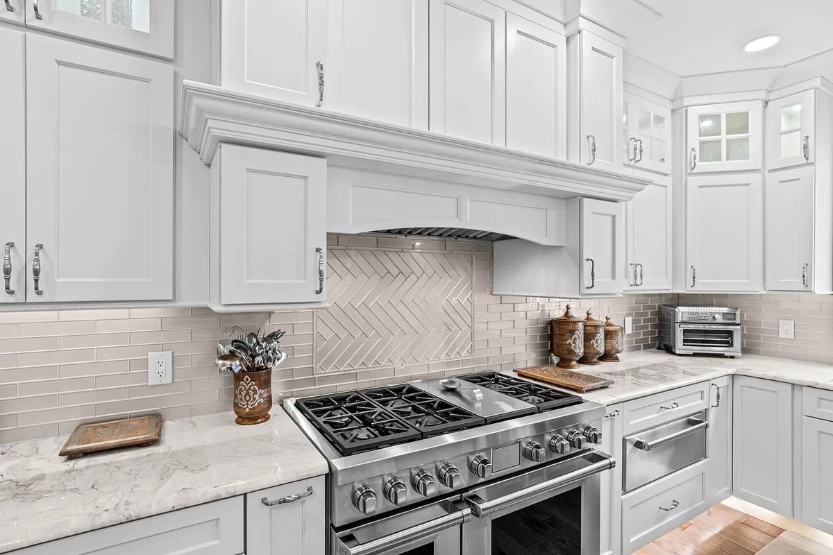 Bright white kitchen with stainless steel stove, ornate range hood, gray backsplash, and countertop decor
