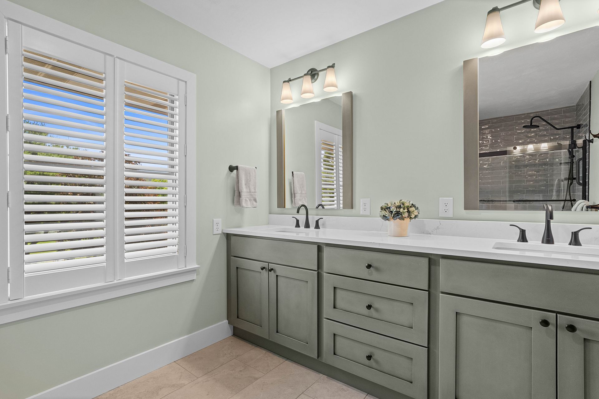 Bright bathroom with sage green vanity, double sinks, mirrors, and white plantation shutters