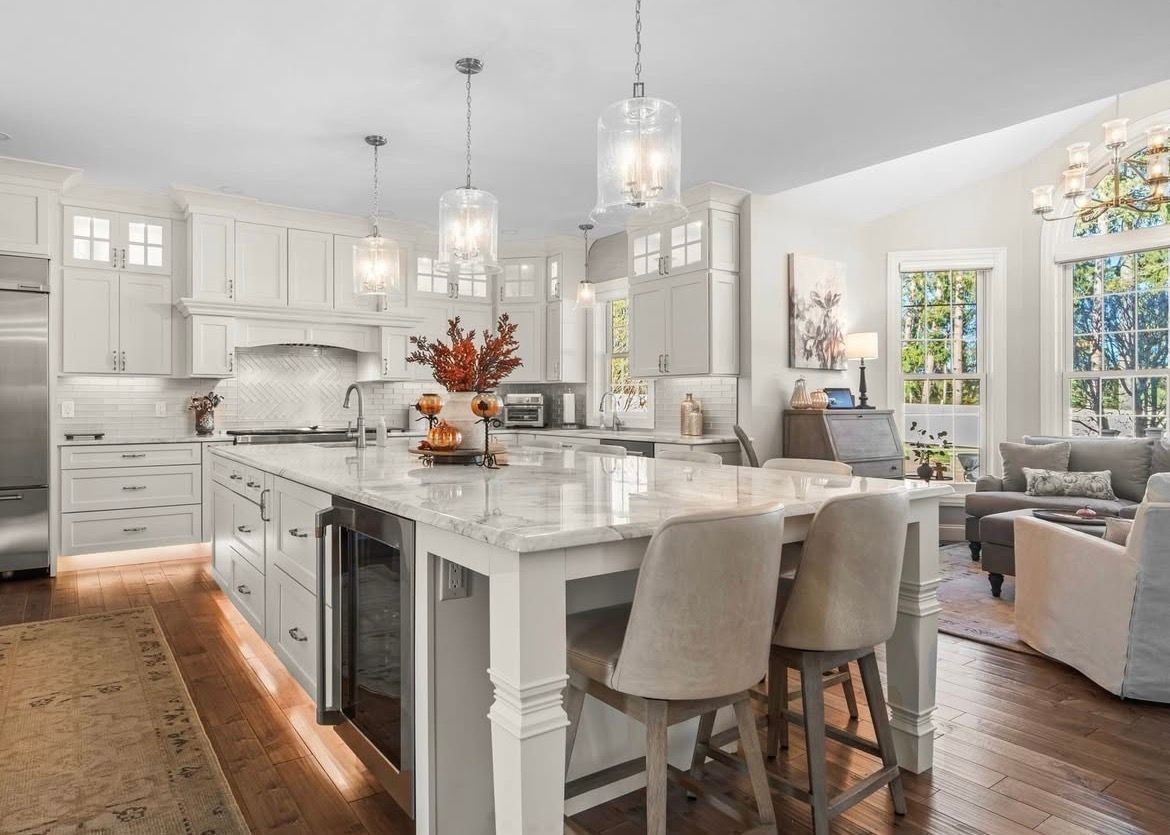 Bright white modern kitchen with island, pendant lights, and hardwood floors, opening to a cozy living area.