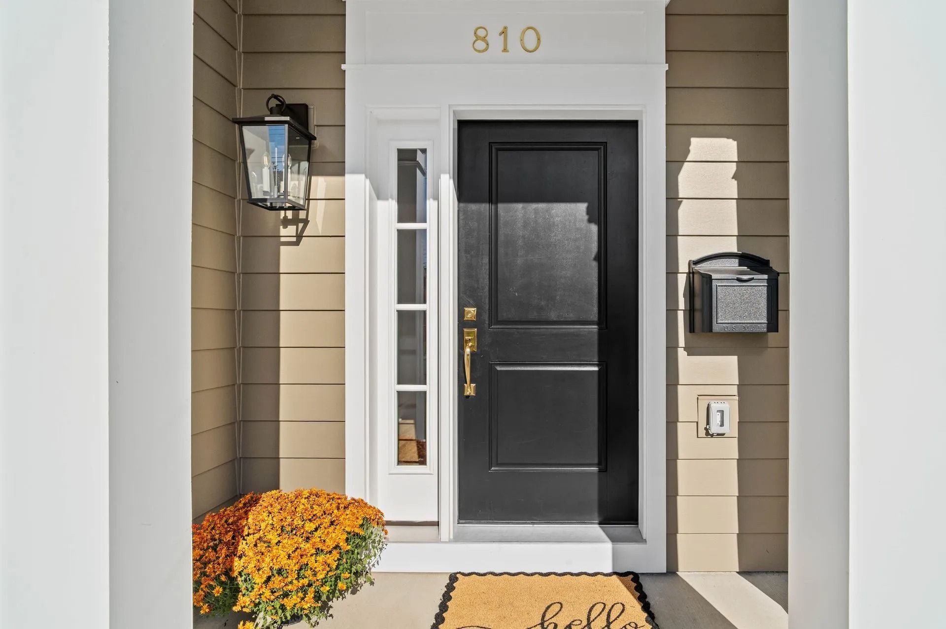 Front porch with black front door, tan siding, wall lantern, mailbox, and orange flowers