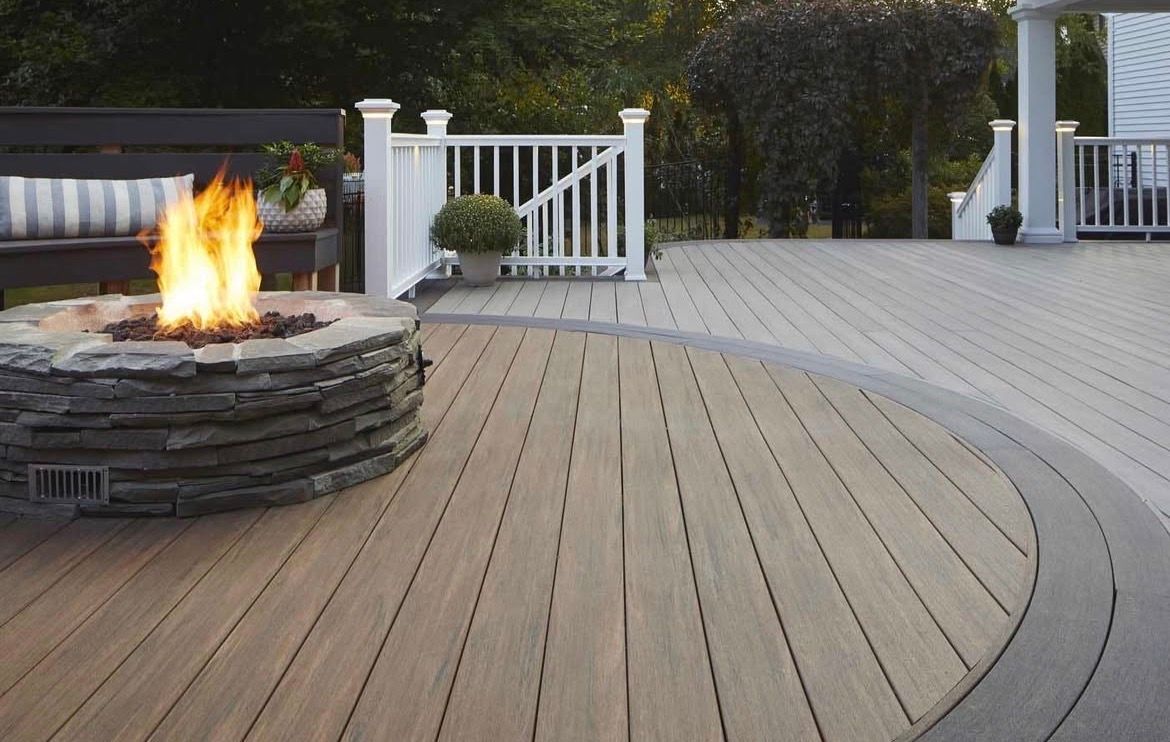 Curved wooden deck with a stone fire pit burning beside a white fence and bench in a backyard