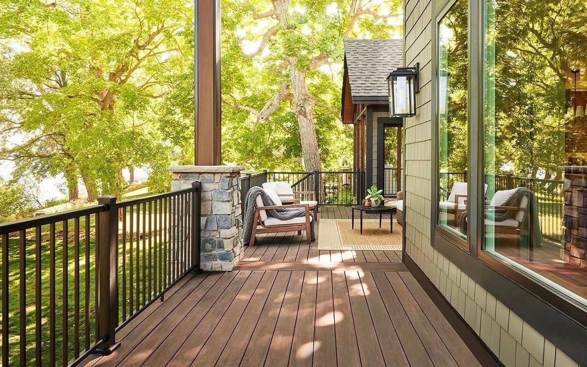 Wooden porch with black railing, cushioned chairs, and leafy trees beside a house exterior