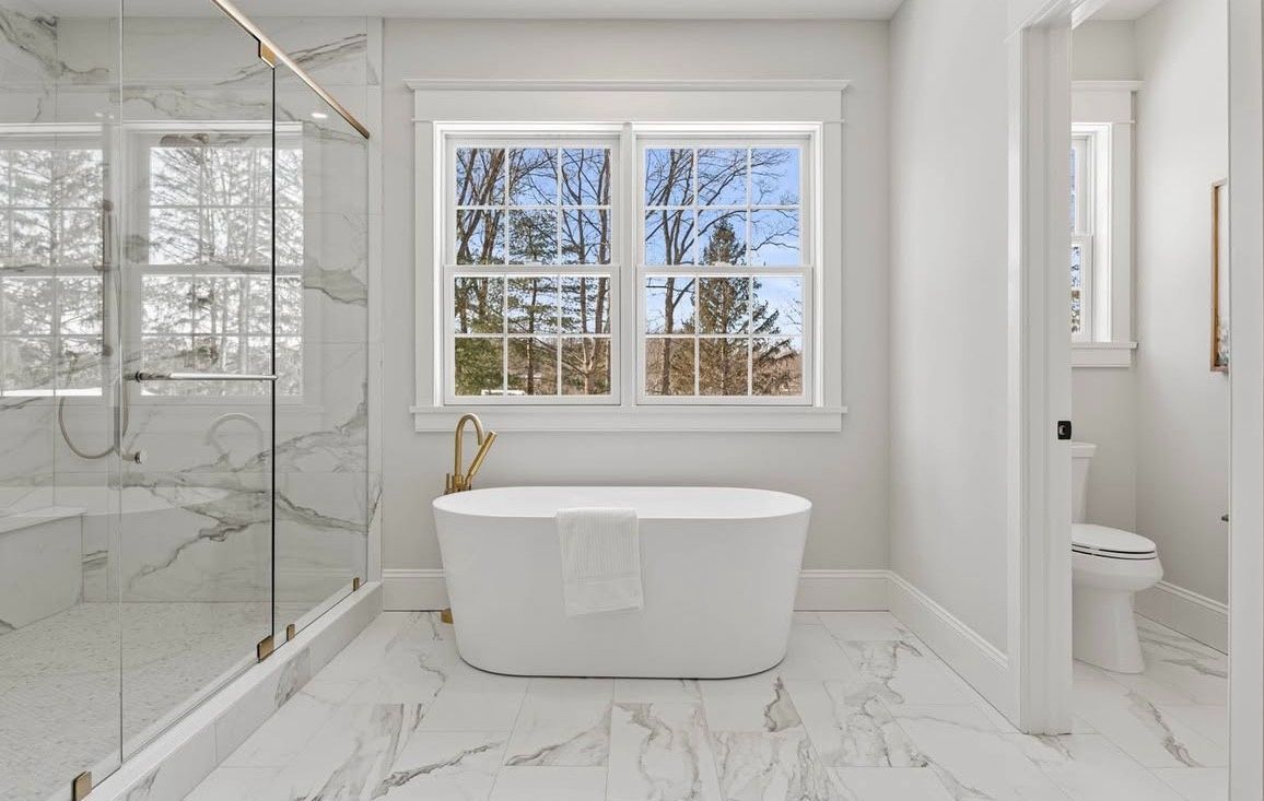 Bright white bathroom with freestanding tub, marble tile, glass shower, and window view of snowy trees