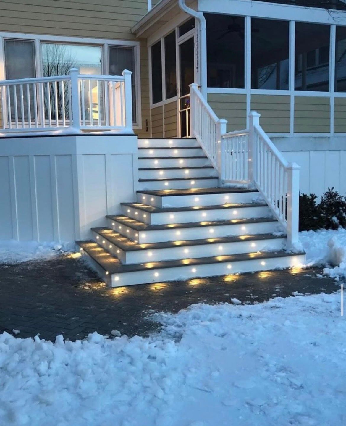 Snowy porch stairs with white railings and warm step lights leading to a house entrance