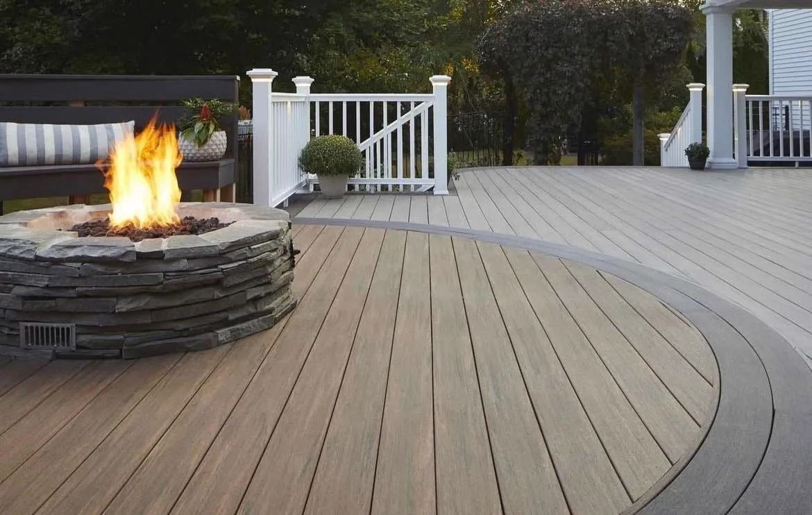 Curved wooden deck with a stone fire pit burning beside white railings and hedges