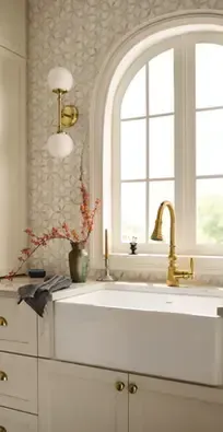 Bright kitchen sink beneath an arched window, with gold faucet and white cabinets