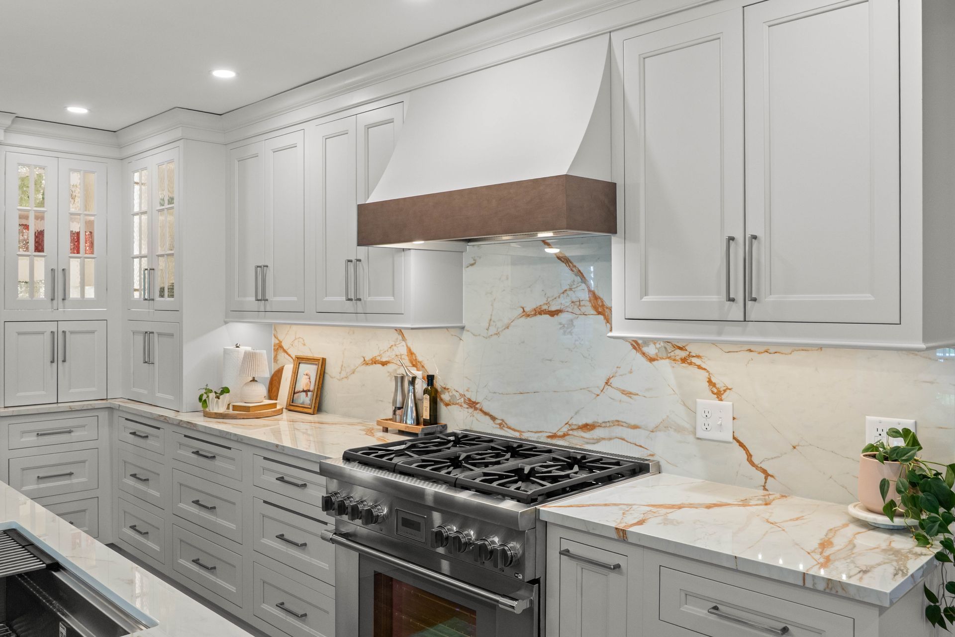 Bright white modern kitchen with gray cabinets, marble countertops, and a gas range under a stainless hood.