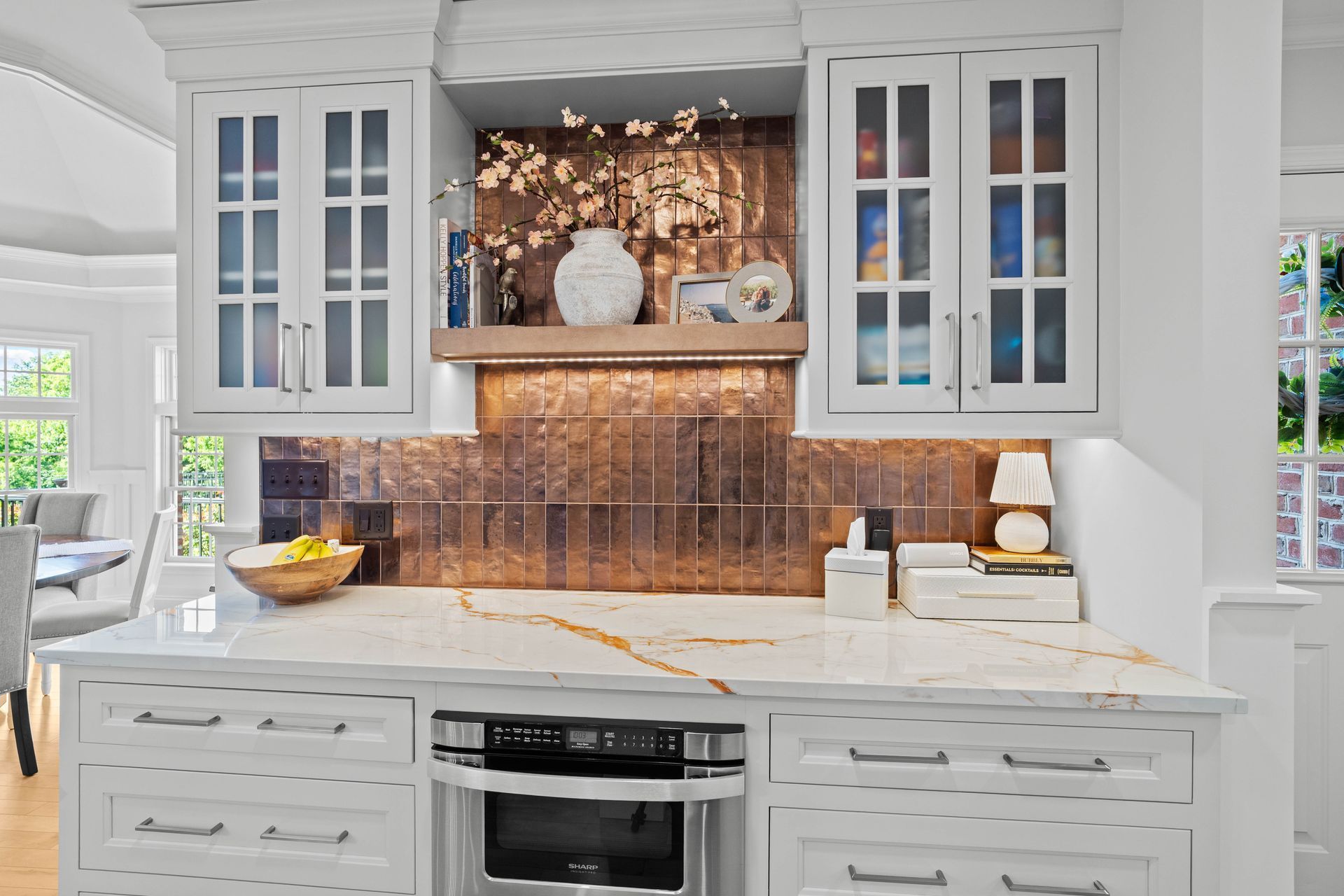 Bright white kitchen with marble counters, glass cabinets, and copper backsplash above a stove