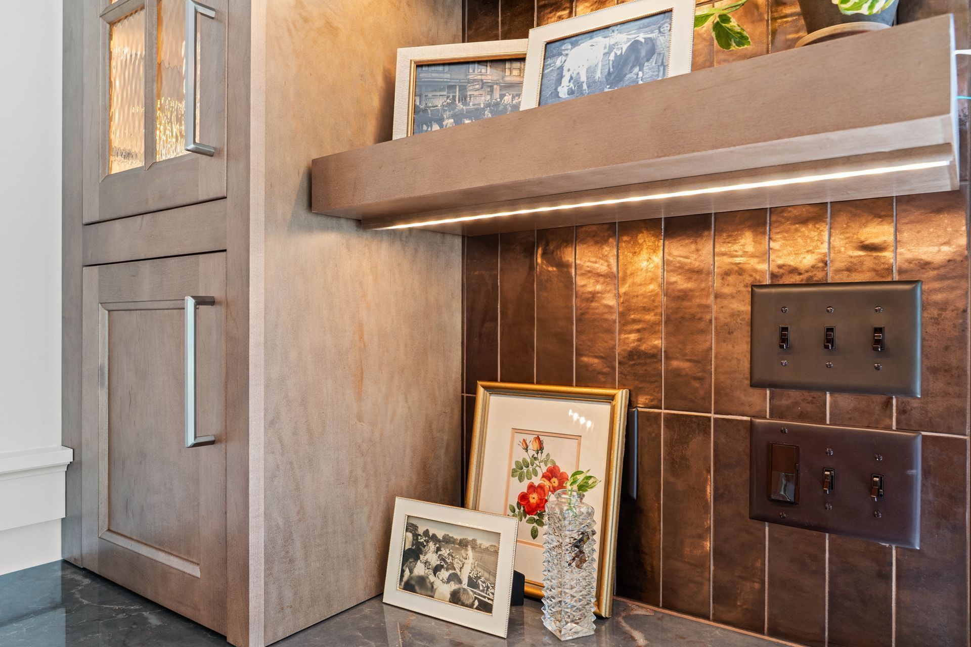 Built-in wooden cabinet nook with framed art, shelf decor, and wall outlet above a dark tile backsplash