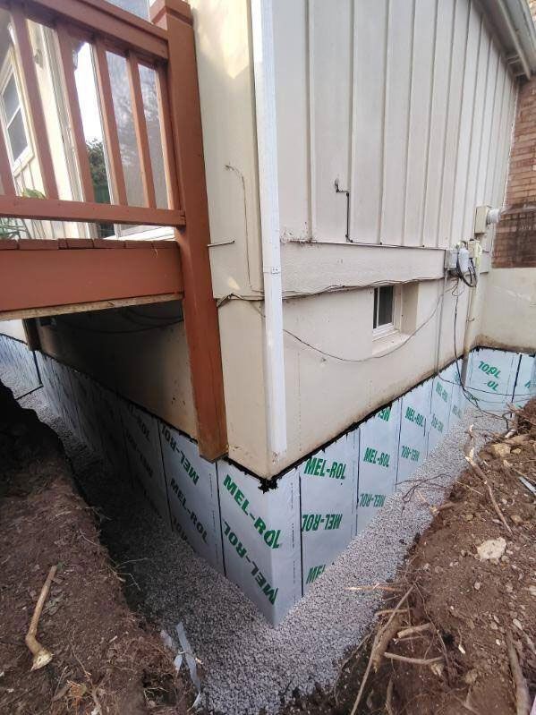 Exterior view of a home's foundation with waterproofing membrane installed, gravel base, and adjacent deck.
