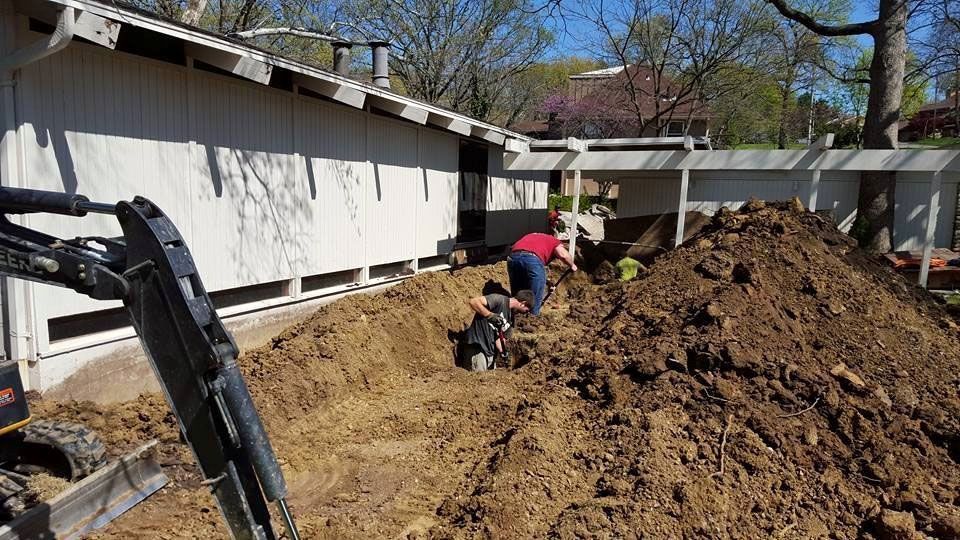 Construction workers excavating dirt near a white building, sunny day.