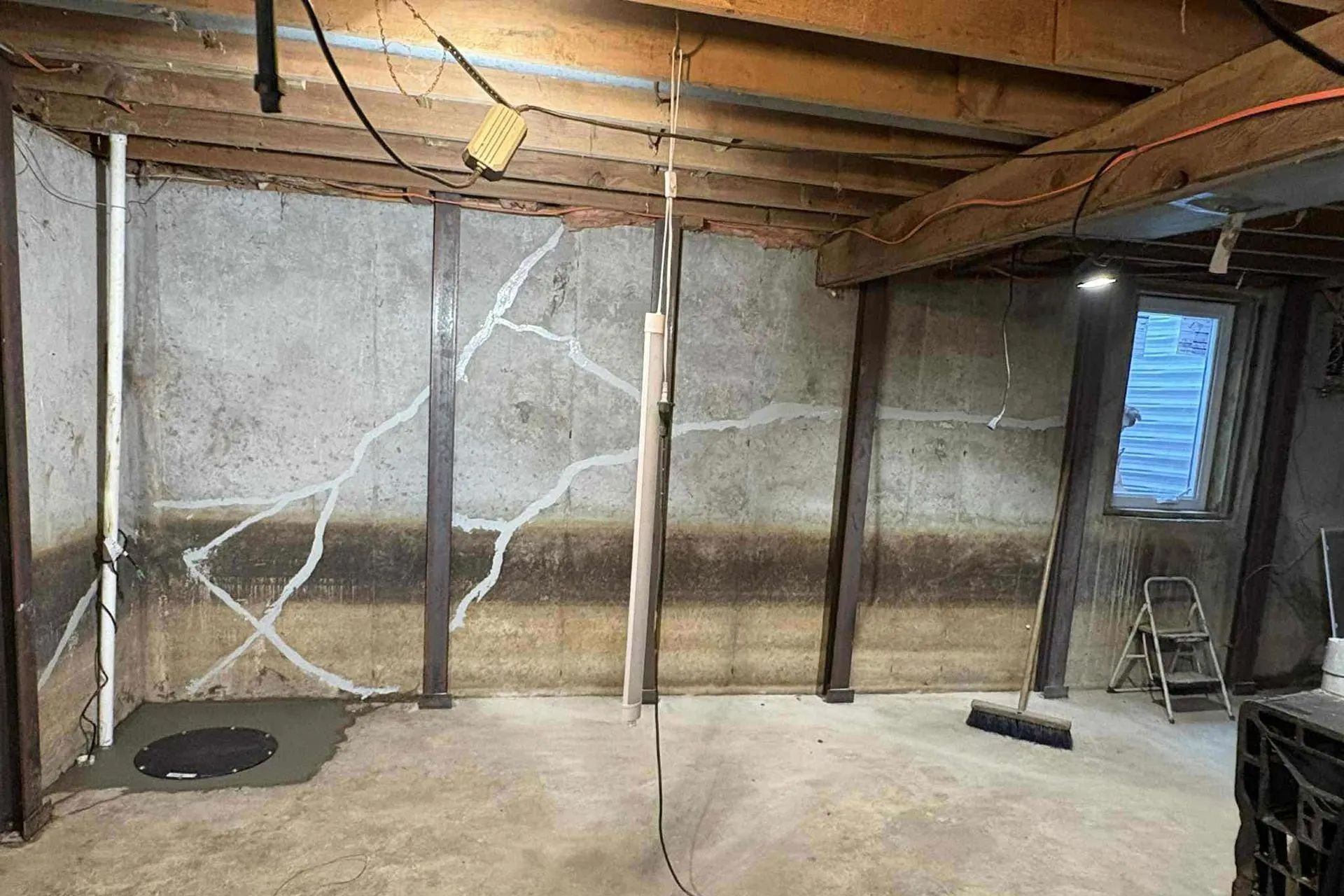 A basement concrete wall with prominent diagonal cracks reinforced by vertical steel support beams.