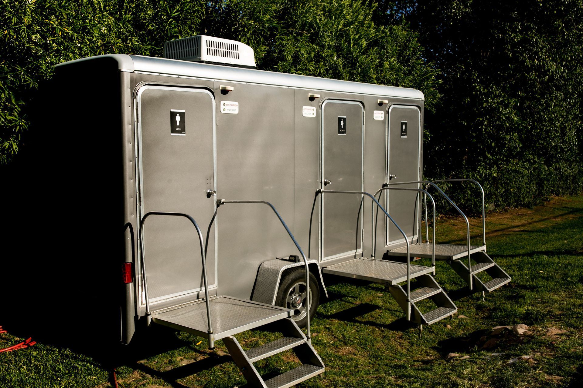 Two silver portable restroom trailers with steps on a grassy lawn beside trees.