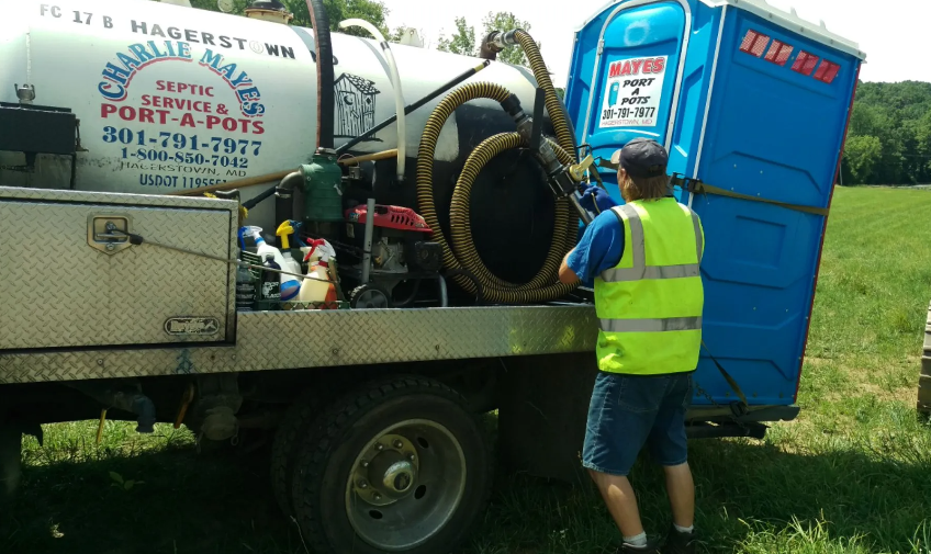 A man in a safety vest services a portable toilet. A truck with a tank and hoses is visible in a field.