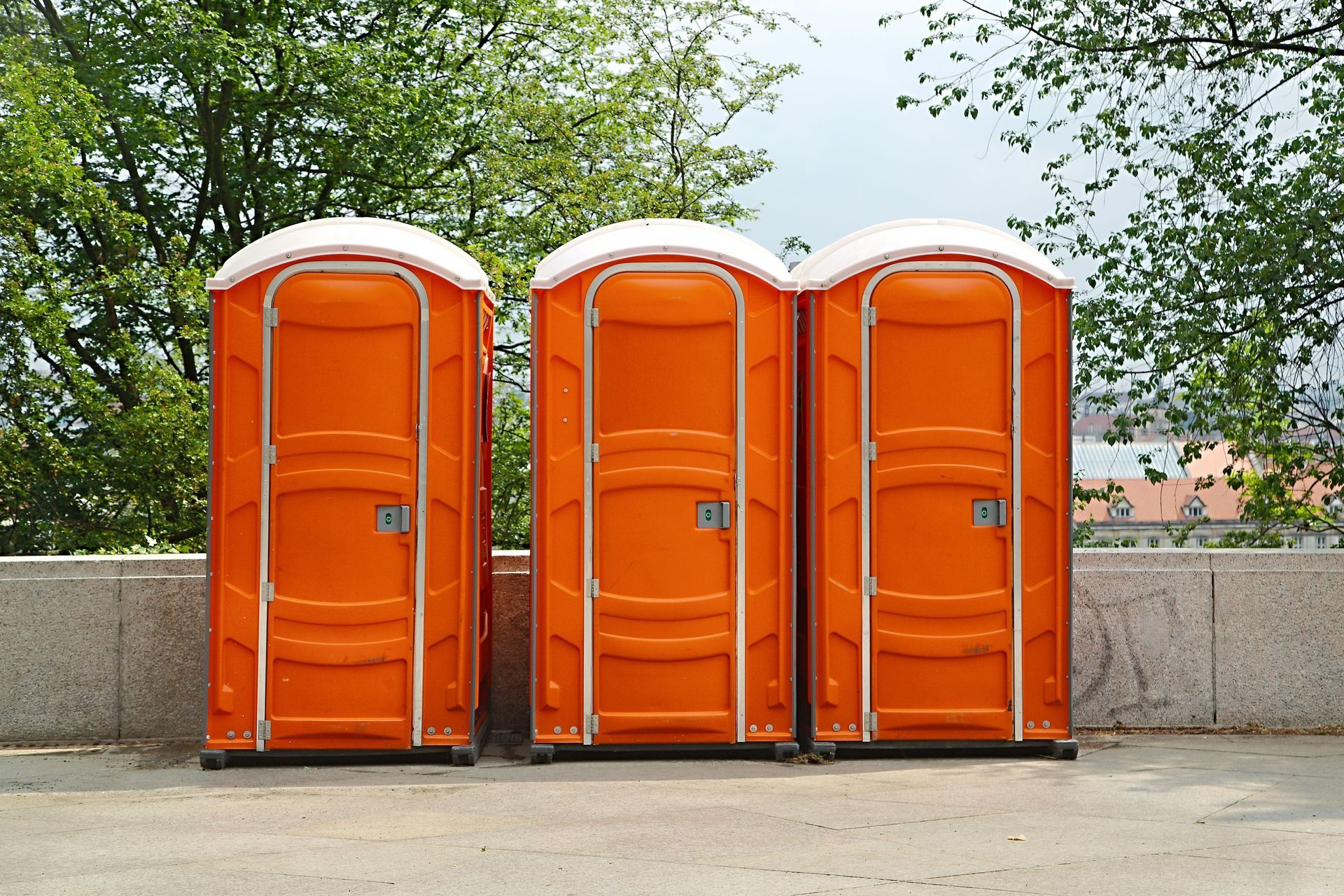 Three orange portable toilets lined up outdoors beside a concrete wall and trees.
