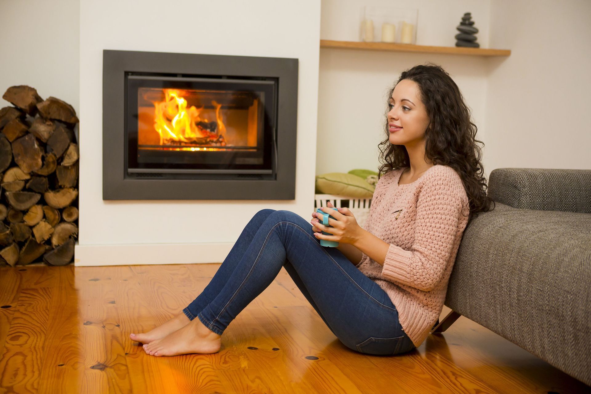 Woman sitting by a fireplace, holding a mug, leaning against a couch, and enjoying the fire.